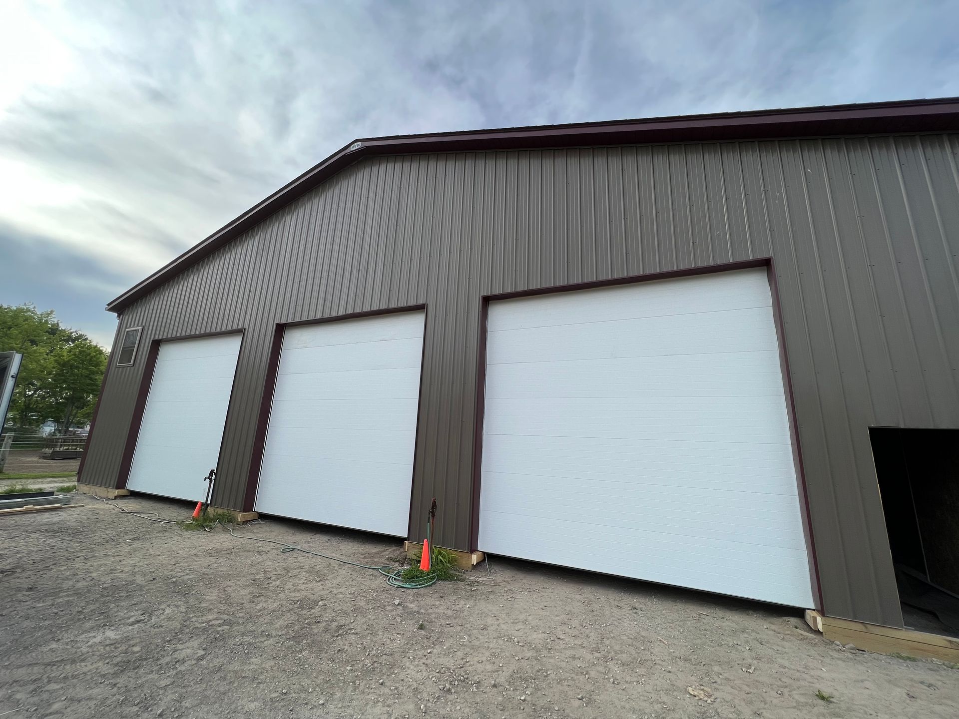 Three white garage doors on a brown metal building, viewed from a gravel lot.
