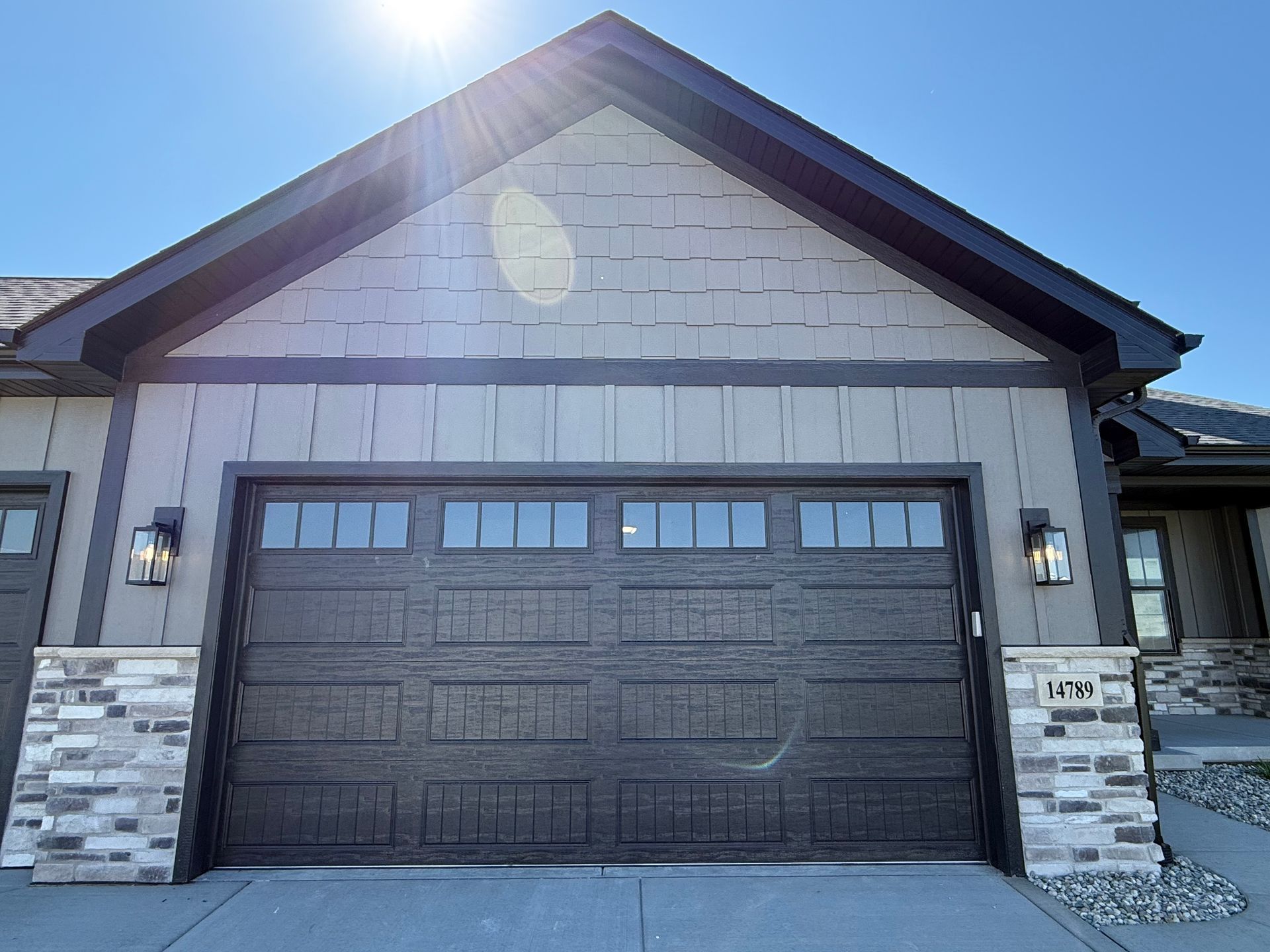 Front view of a modern house with a dark garage door, stone accents, and bright sunlight overhead