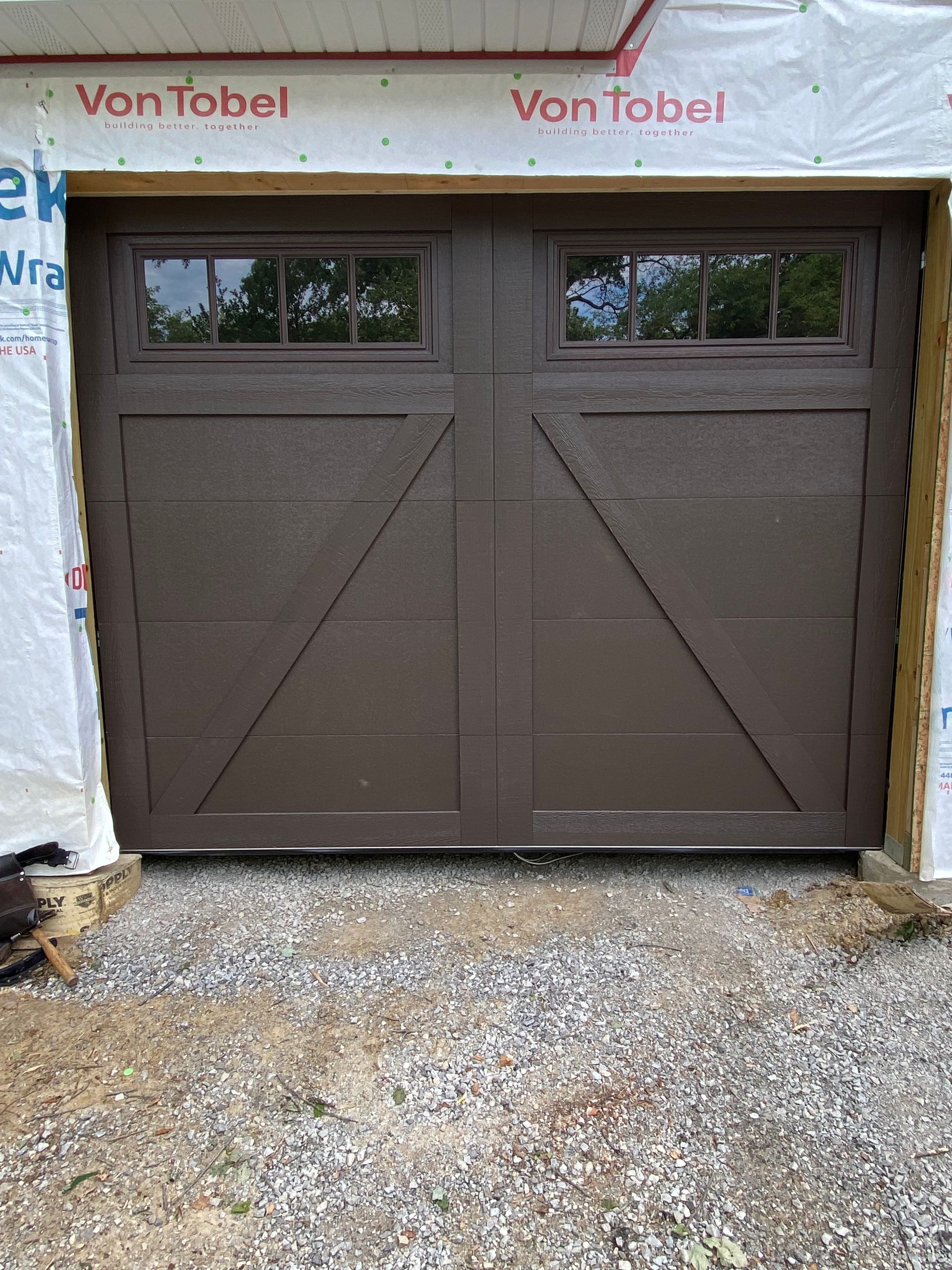 Brown double garage door with small windows, set in unfinished siding above a gravel driveway.