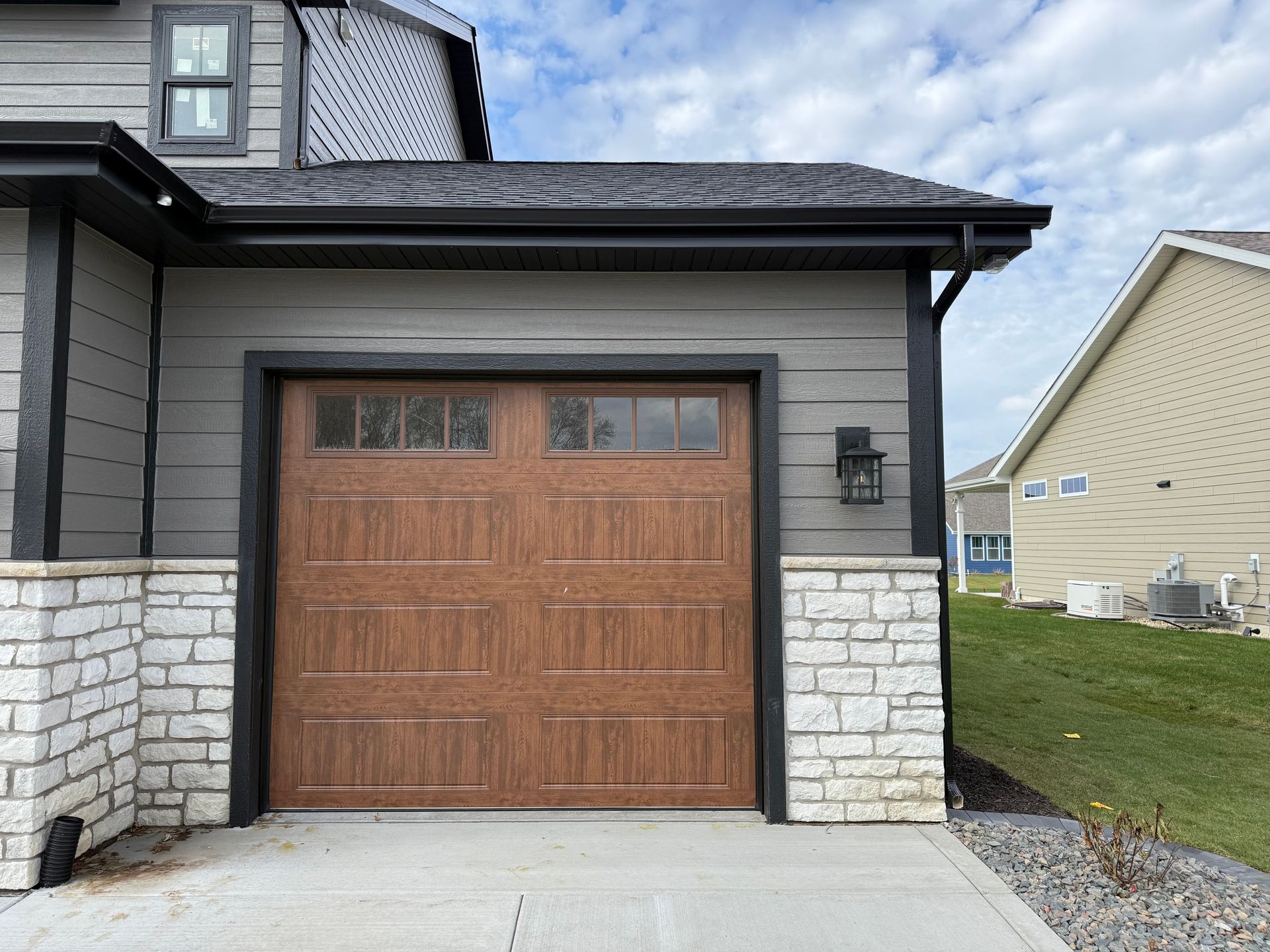 Wooden garage door on a gray house with stone siding and a small driveway, under a cloudy sky
