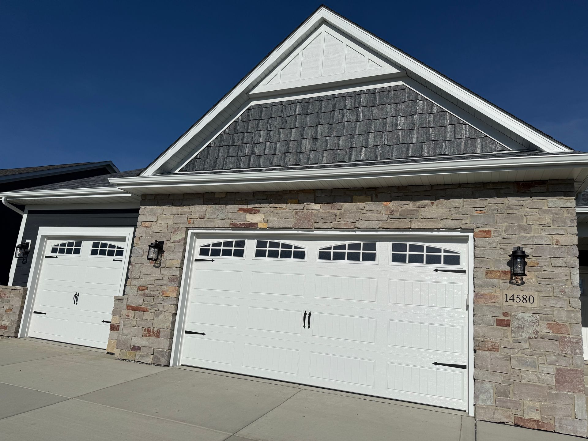 Suburban house exterior with stone facade, two white garage doors, and a gabled roof under a clear blue sky