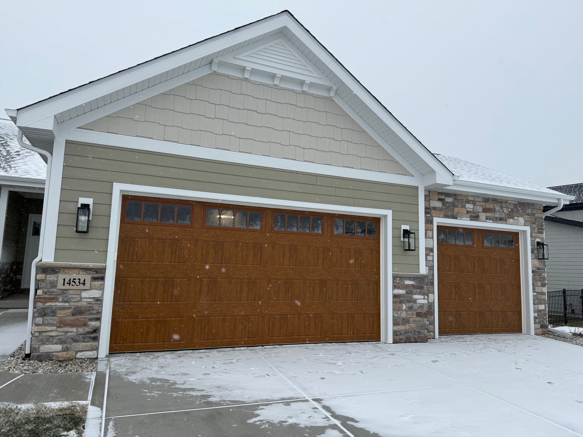 Snow-covered two-car garage with tan siding and wooden doors in a suburban home exterior