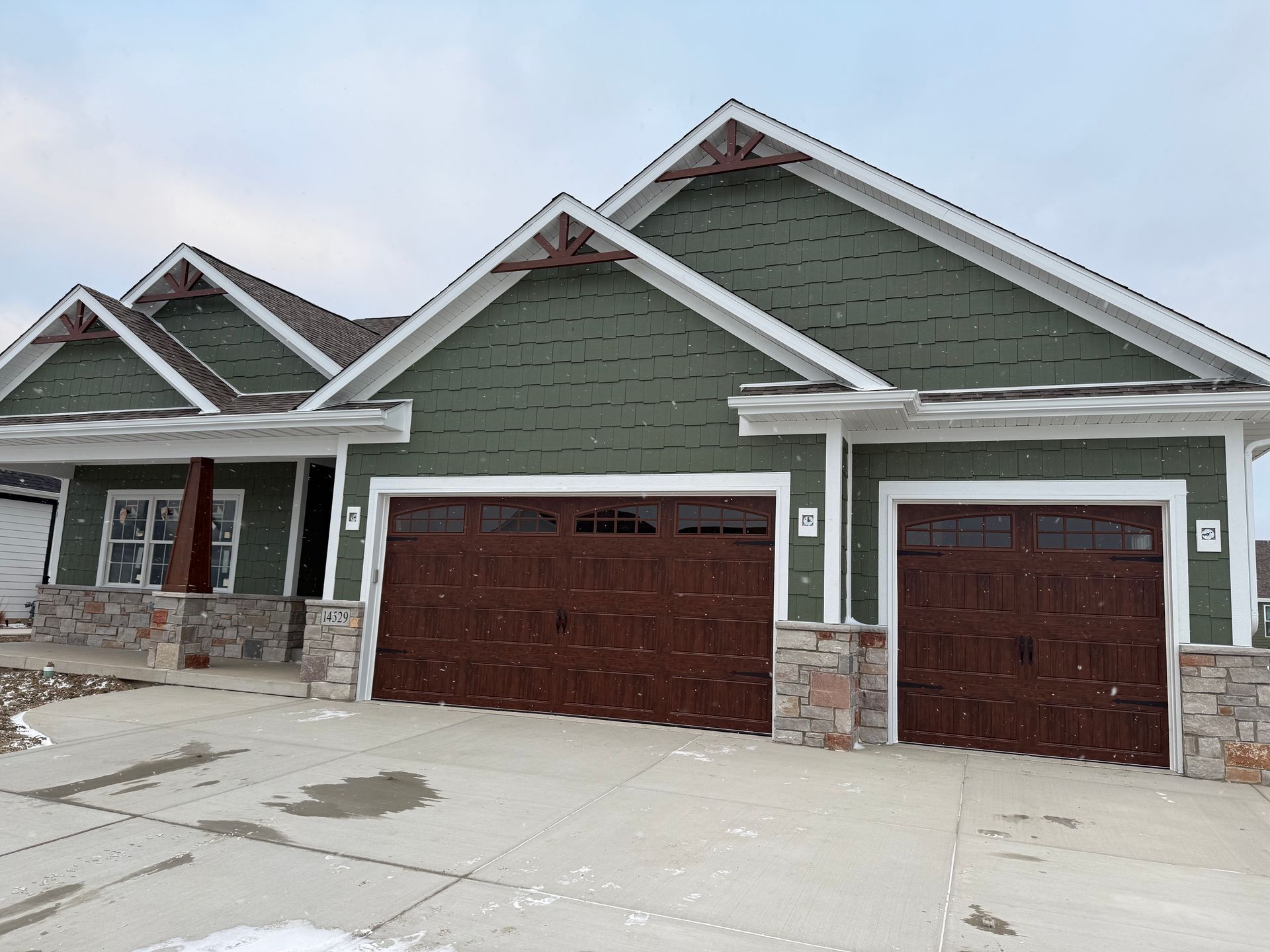 Suburban house exterior with green siding, stone accents, and two brown garage doors in winter.