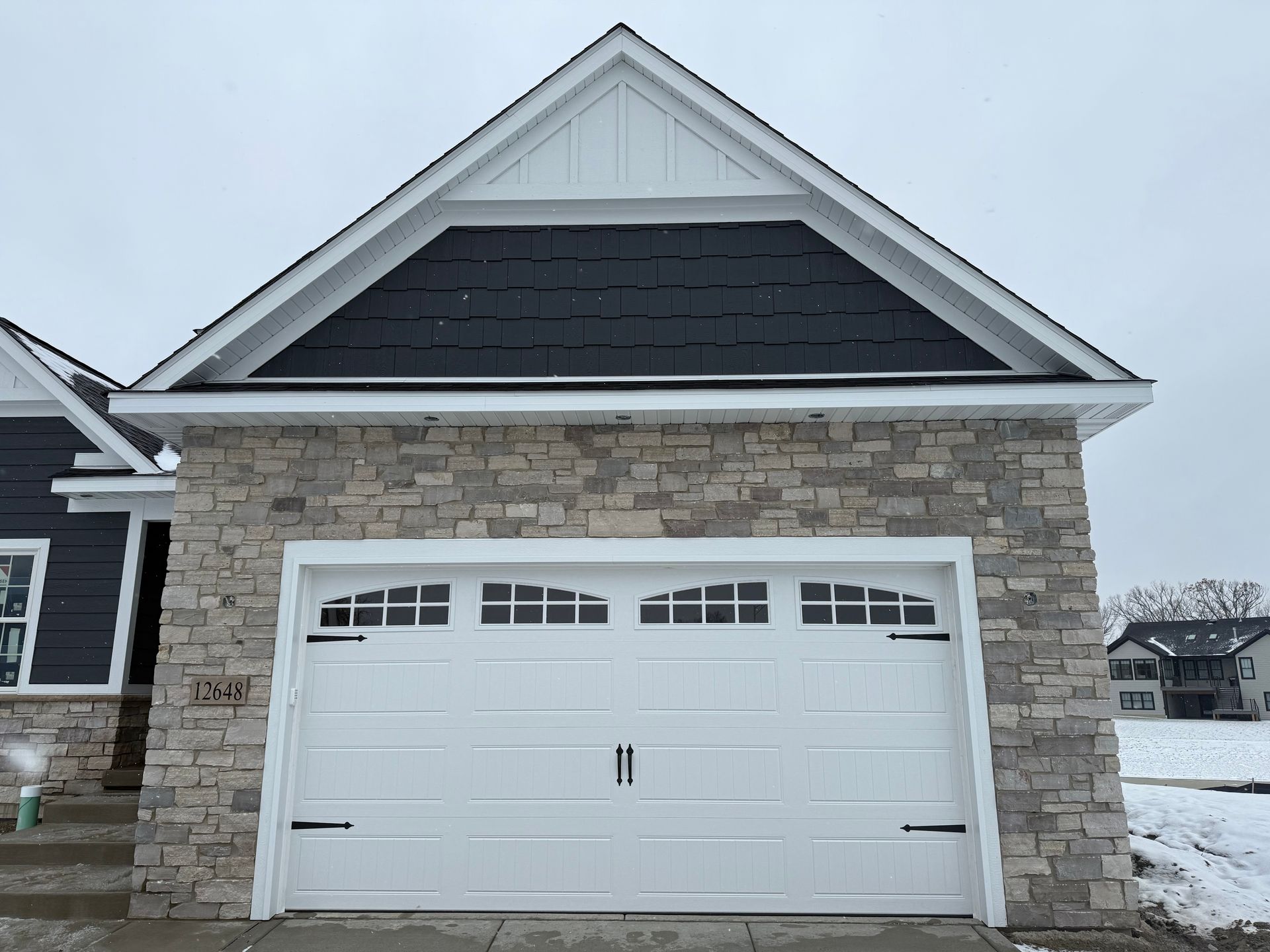 White garage with stone exterior and dark shingled gable roof in a snowy setting