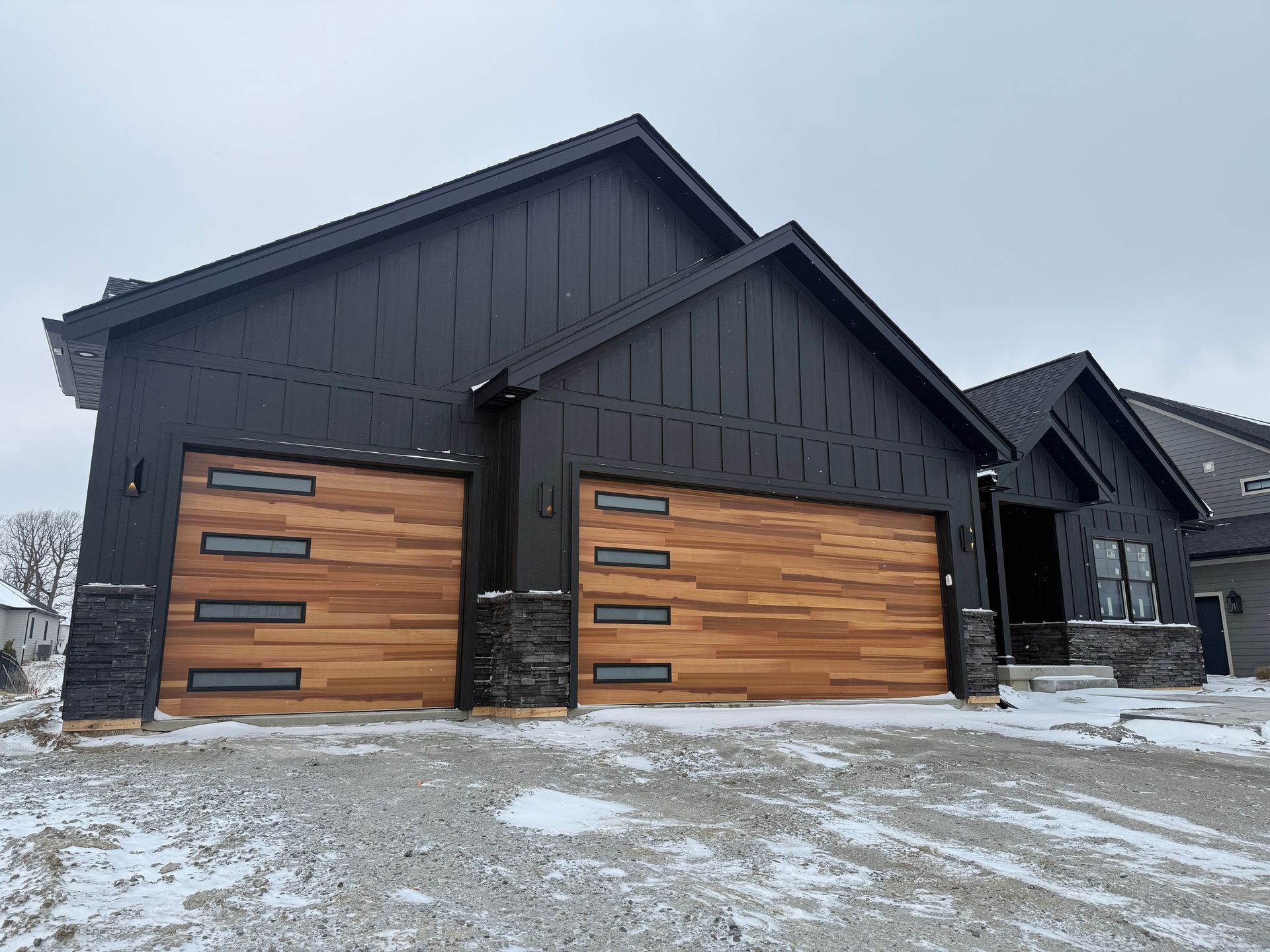 Modern black house exterior with two wood garage doors and snow-covered driveway
