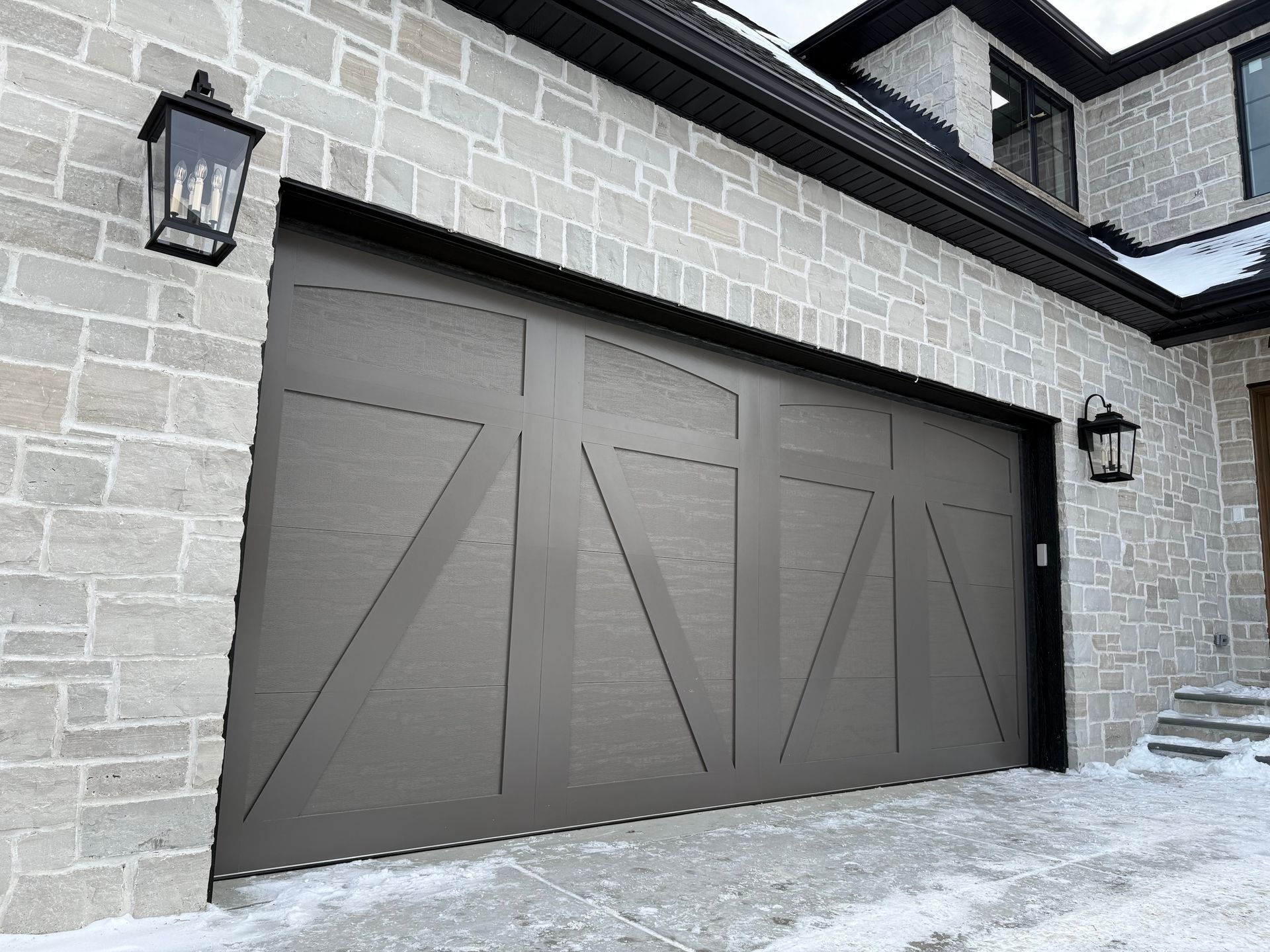 Modern gray garage door on a stone house exterior with black lanterns and snow on the ground