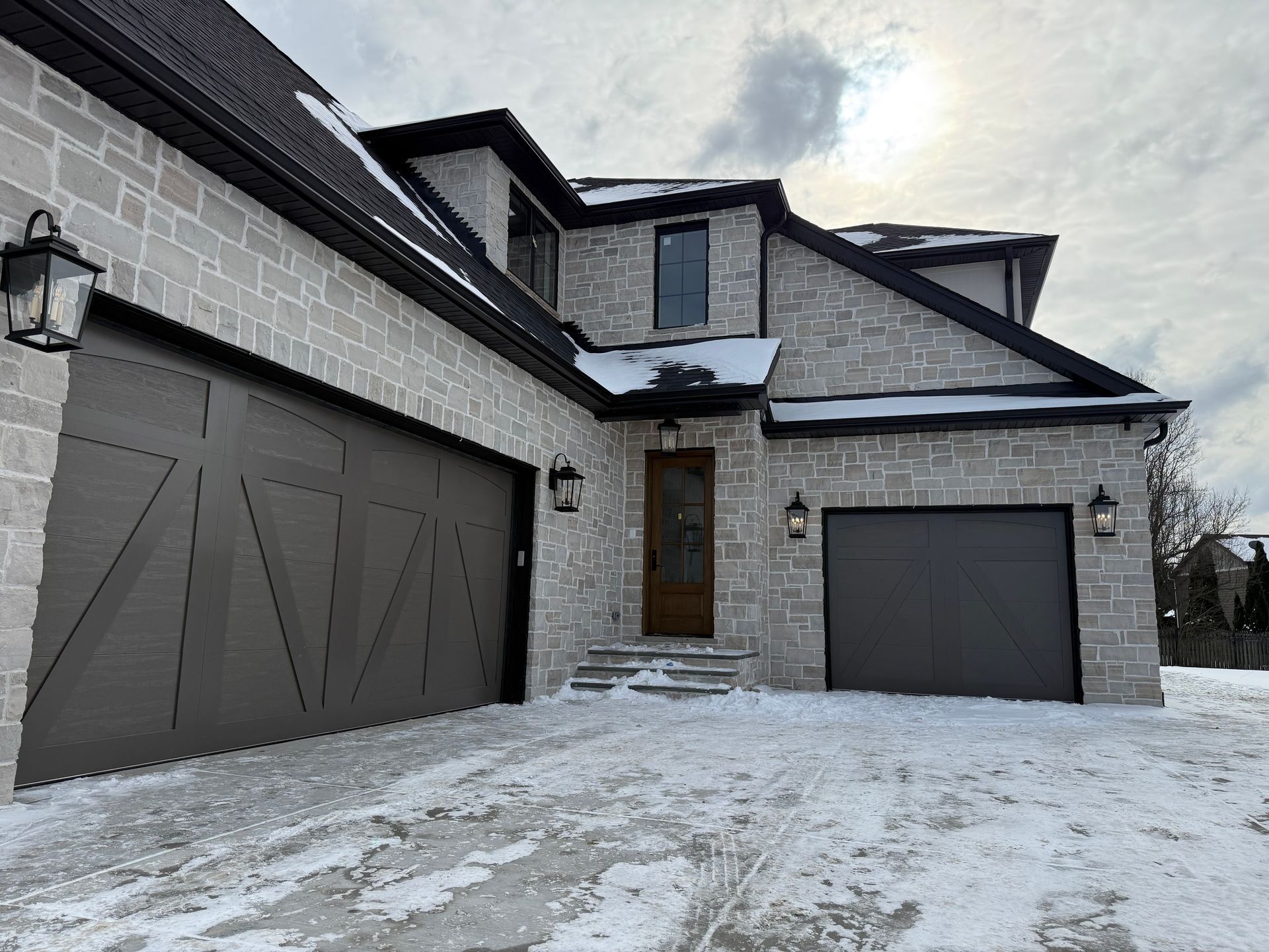 Snow-covered modern house exterior with two garages and a front door under a cloudy sky