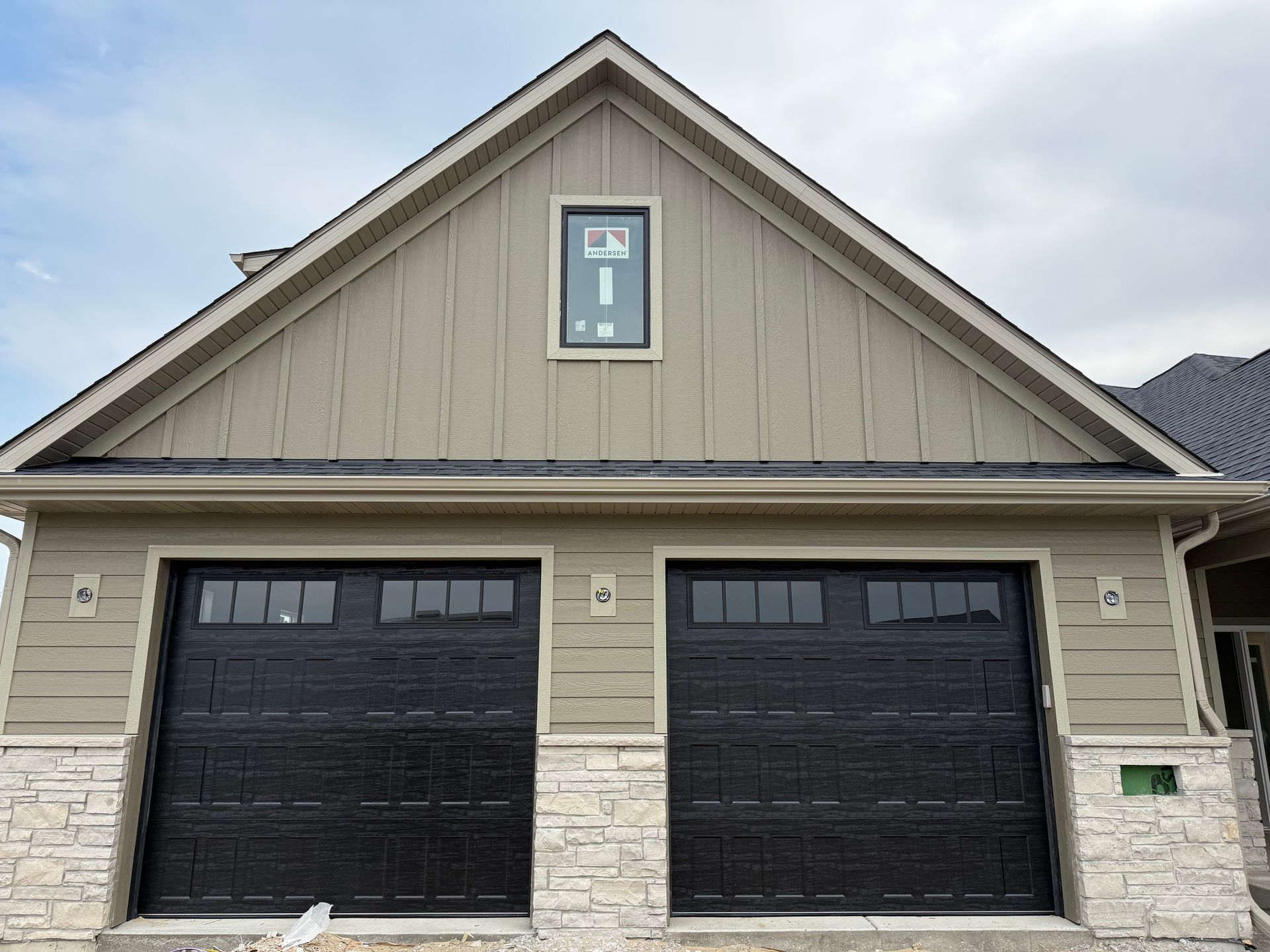 Front view of a beige house with two black garage doors and a peaked roof.