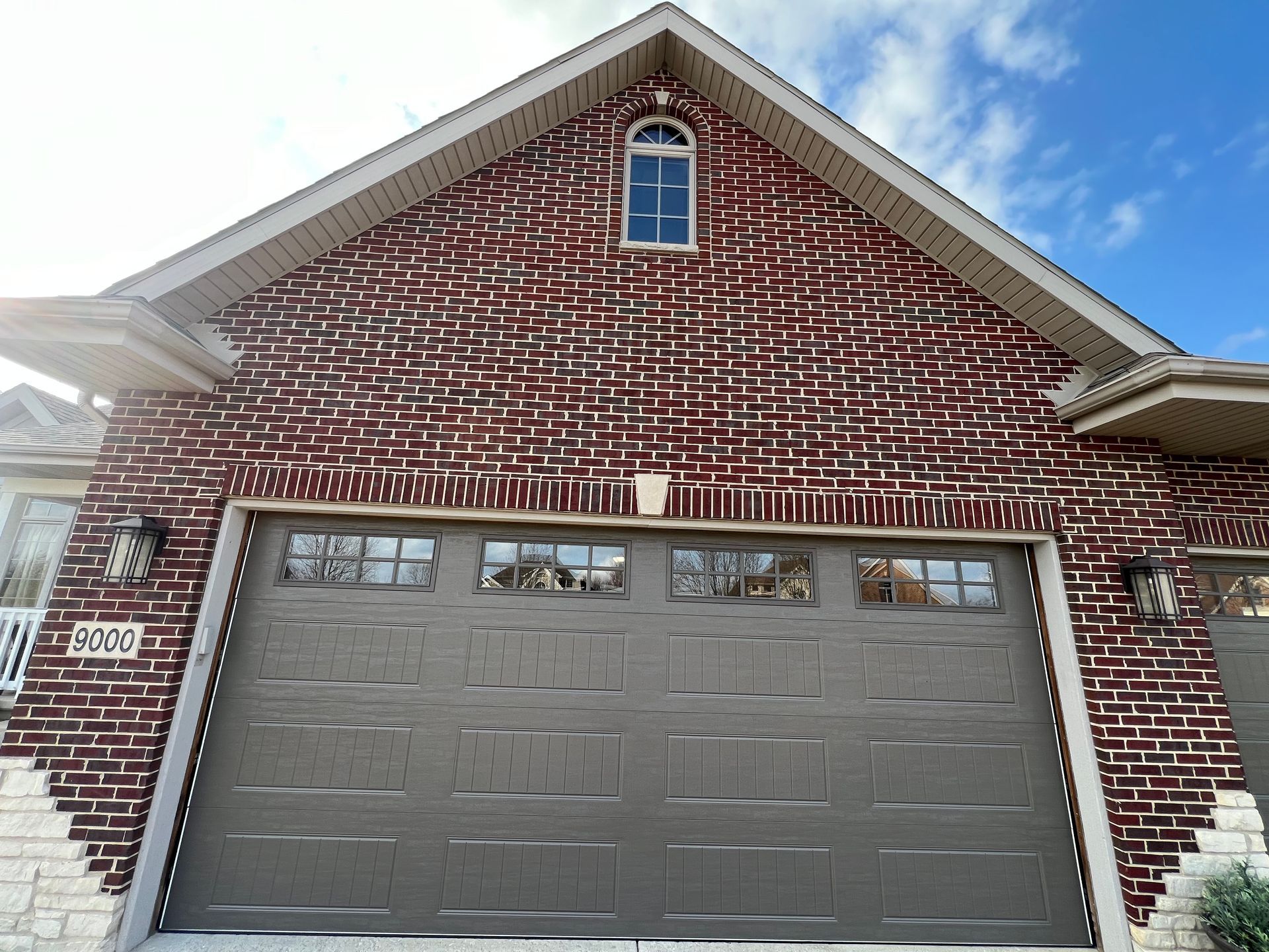 Brick house with a gray two-car garage door and a small arched window above it under a blue sky