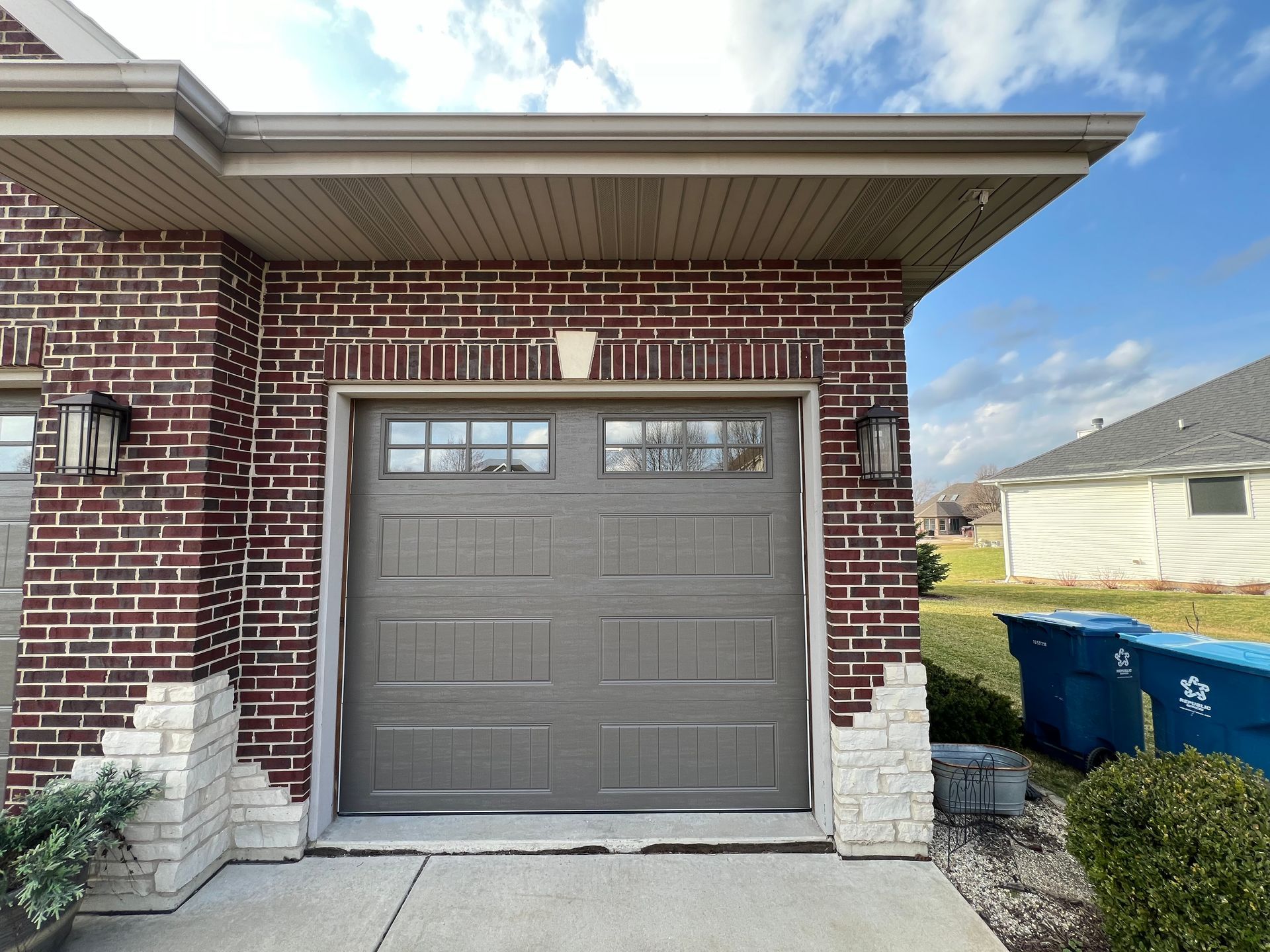 Brick house garage with a gray door, white trim, and blue recycling bins beside it