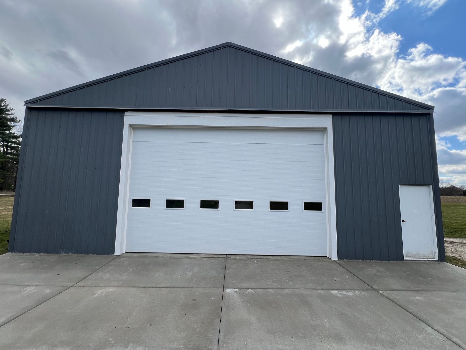 Gray metal warehouse with a white roll-up garage door and a small side door under a cloudy sky.