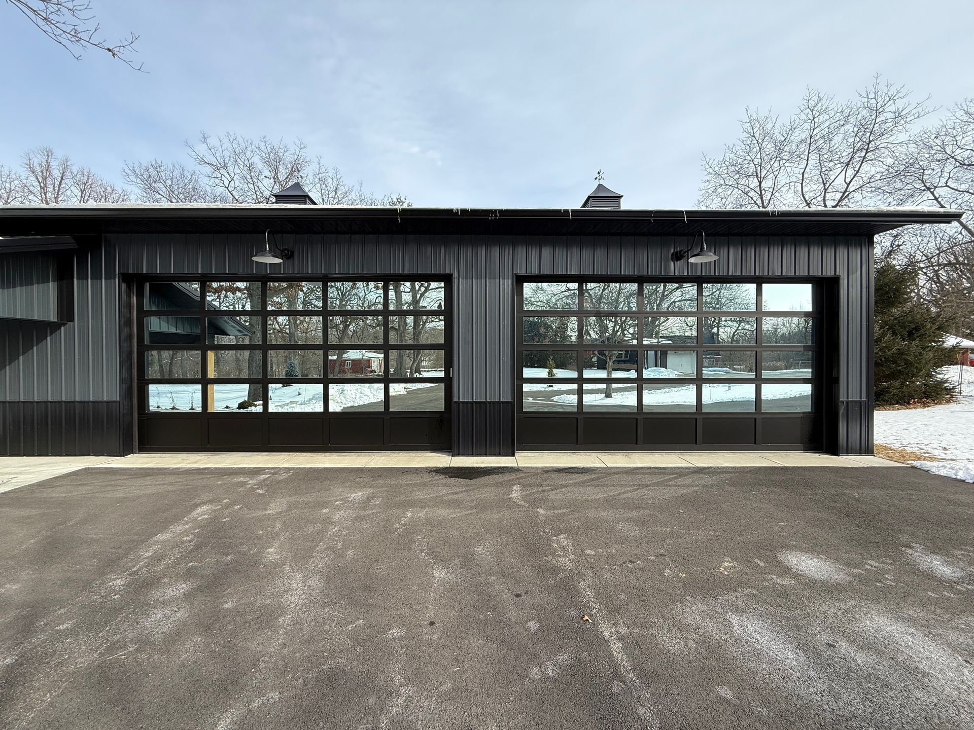 Black garage with two glass-paneled doors and a gravel driveway under a cloudy sky