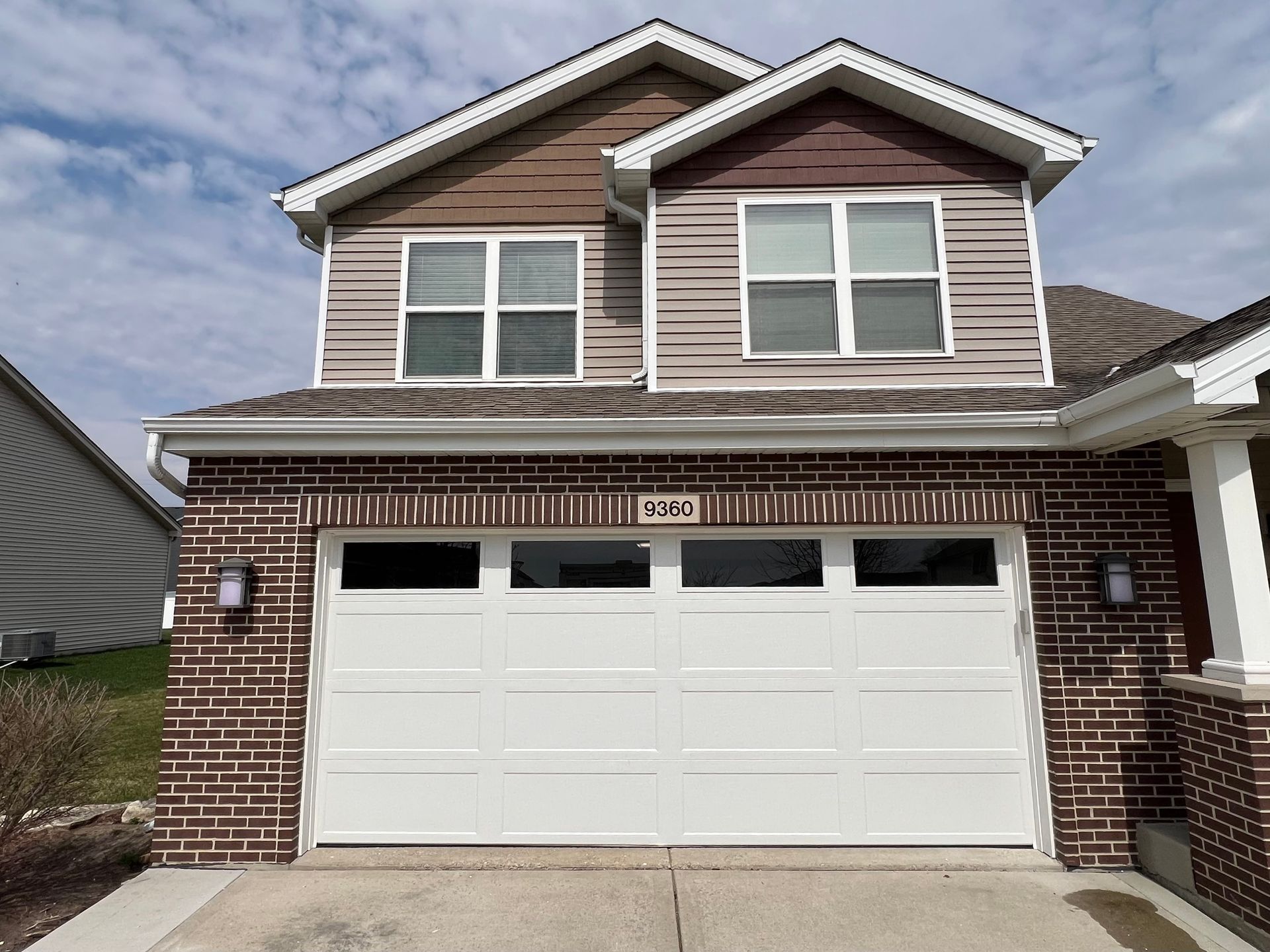 Two-story brick-and-beige house with a white garage door and dormer windows under a cloudy sky