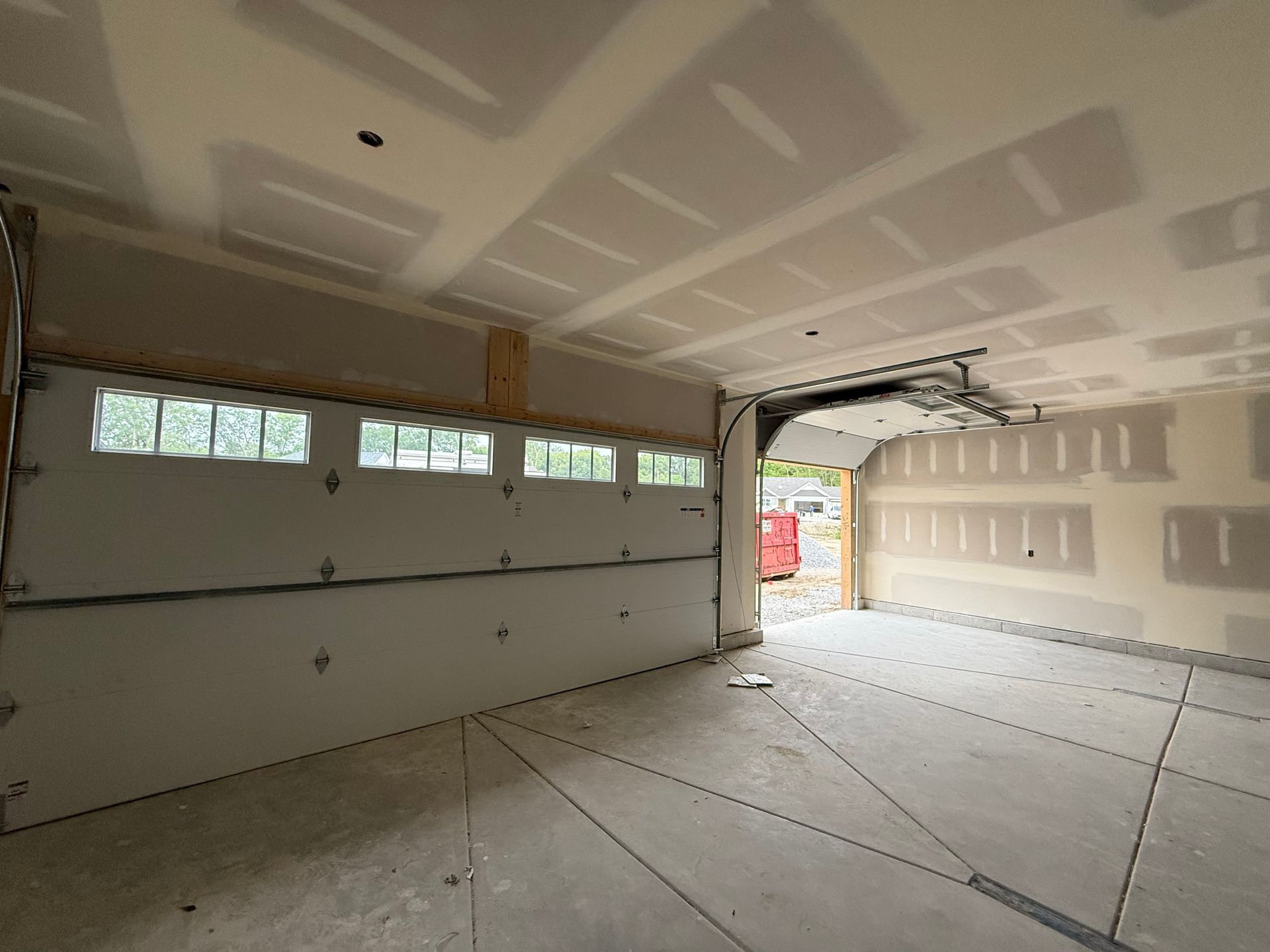 Empty unfinished garage with drywall, exposed door track, and daylight through an open door.