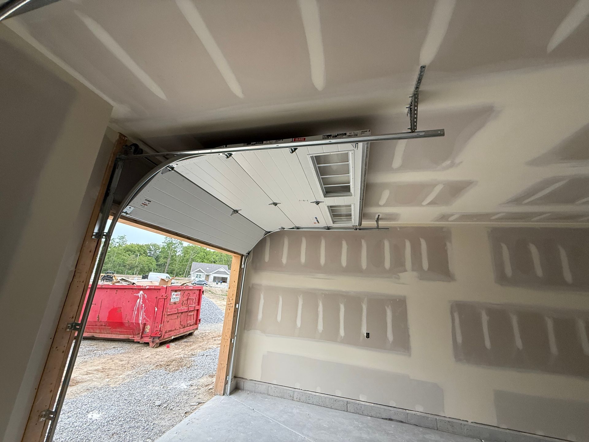 Inside an unfinished garage, looking out through an open door at a red dumpster and gravel outside.