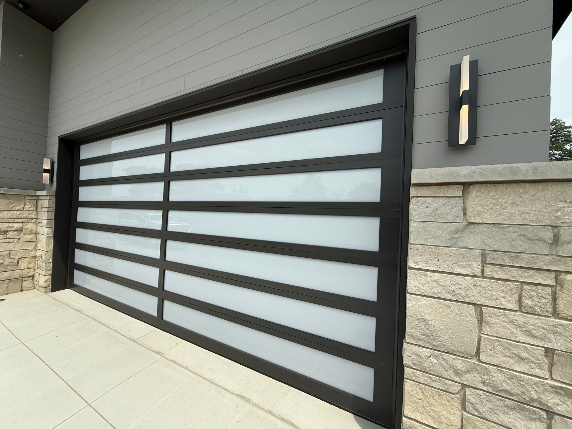Modern frosted-glass garage door with dark horizontal slats beside a stone wall and exterior light fixture