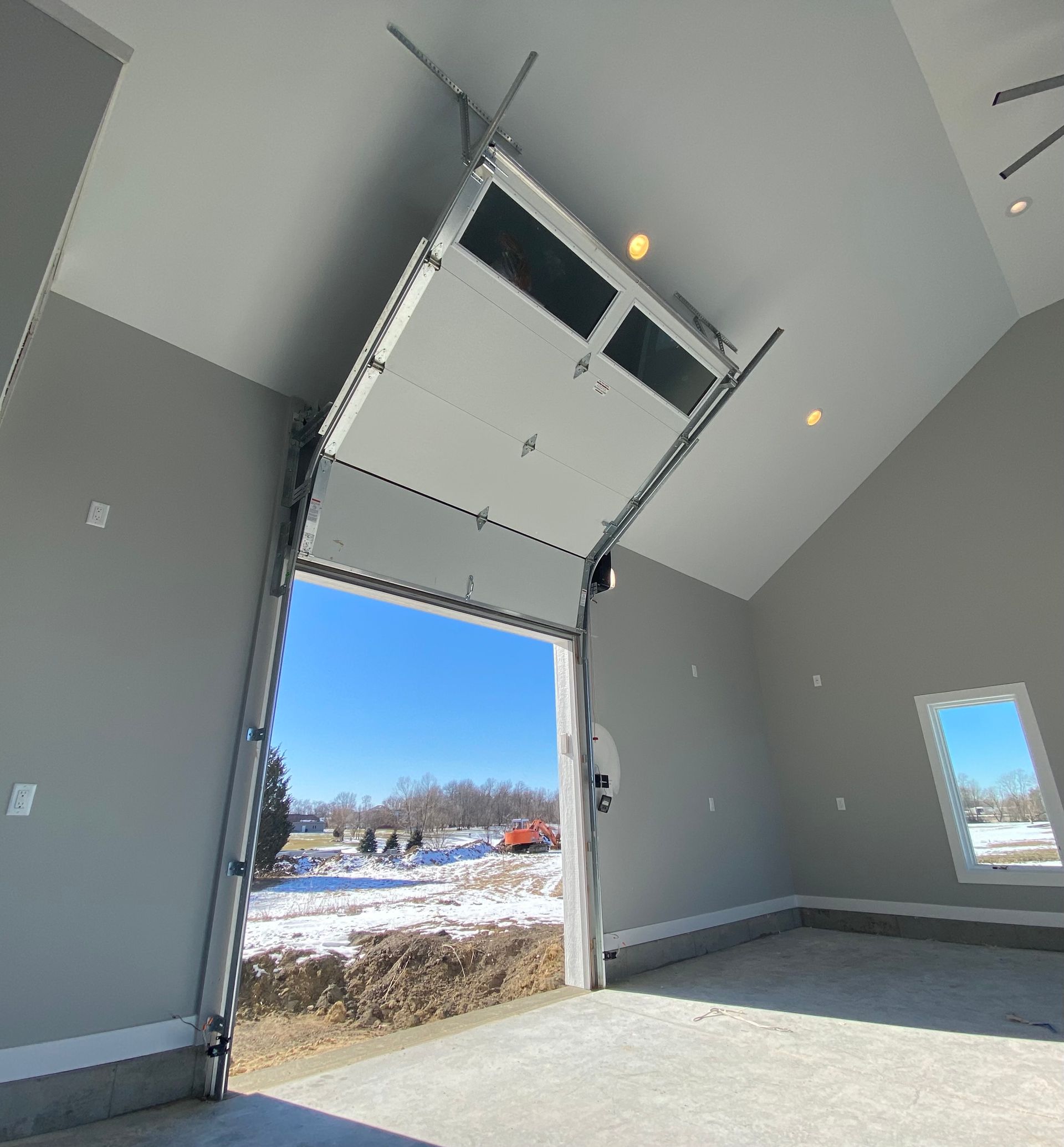 Empty interior with open garage door, white walls, and snowy outdoor view under high ceilings