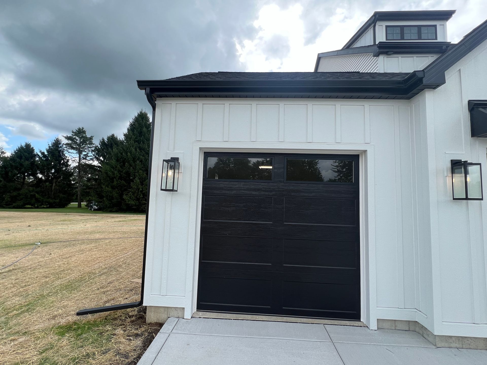 White barn-style garage with a black door and wall lanterns beside a grassy field under a cloudy sky