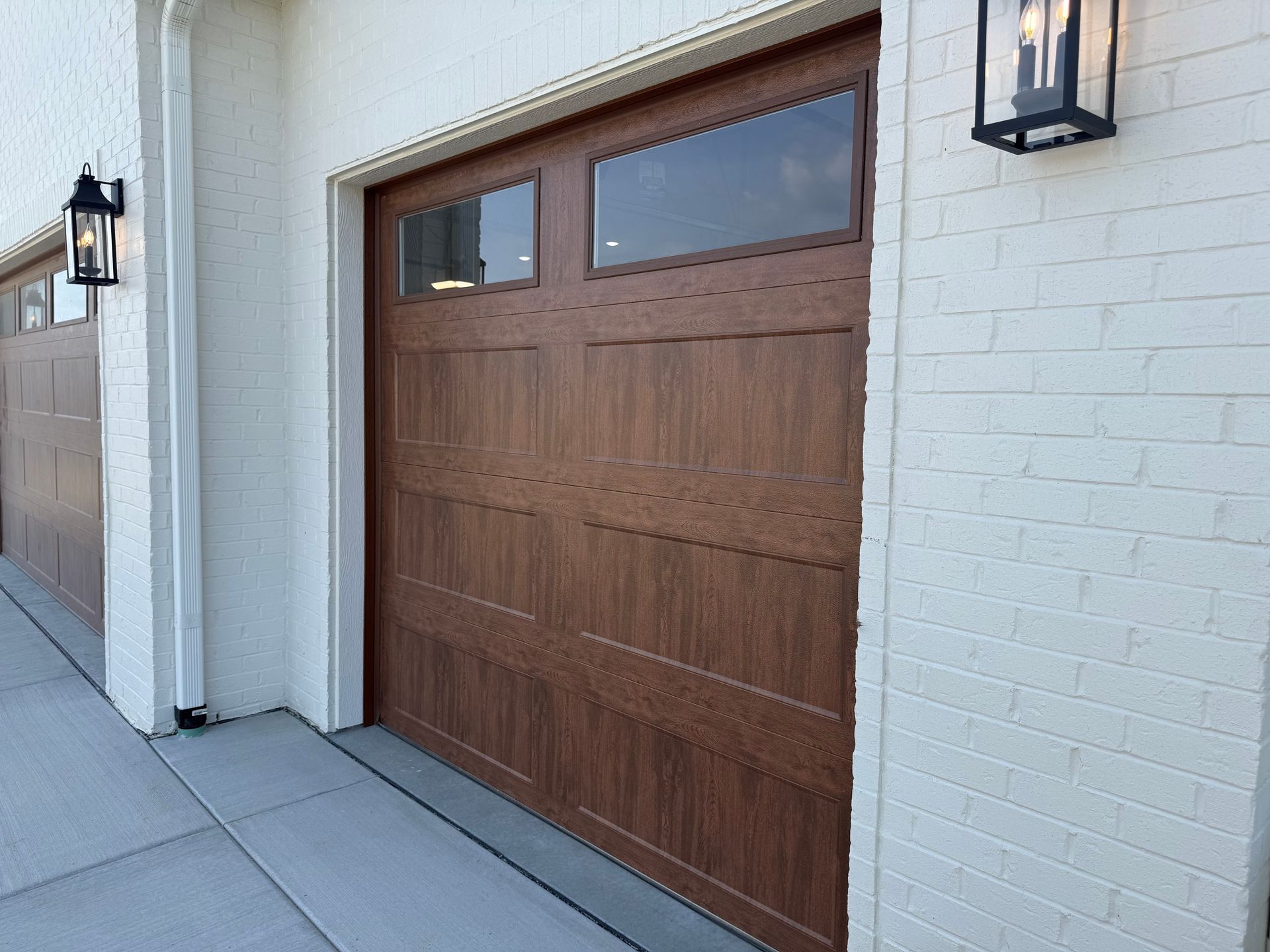 Brown garage door on a white stucco house with wall-mounted lanterns and a concrete driveway.
