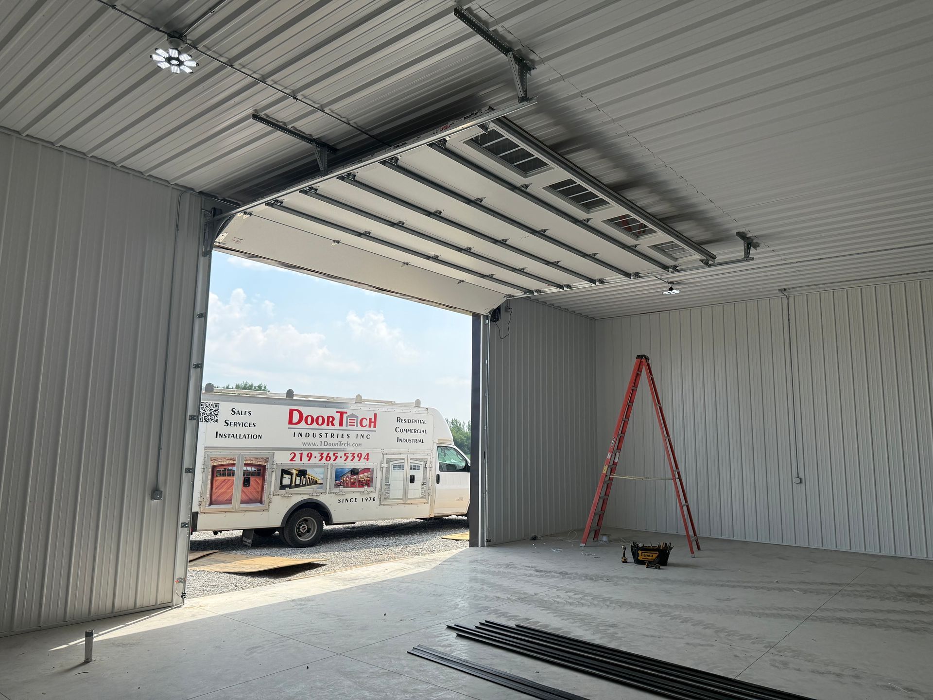 Inside a metal garage bay, a white service truck is parked outside; a red ladder and tools sit on the floor.