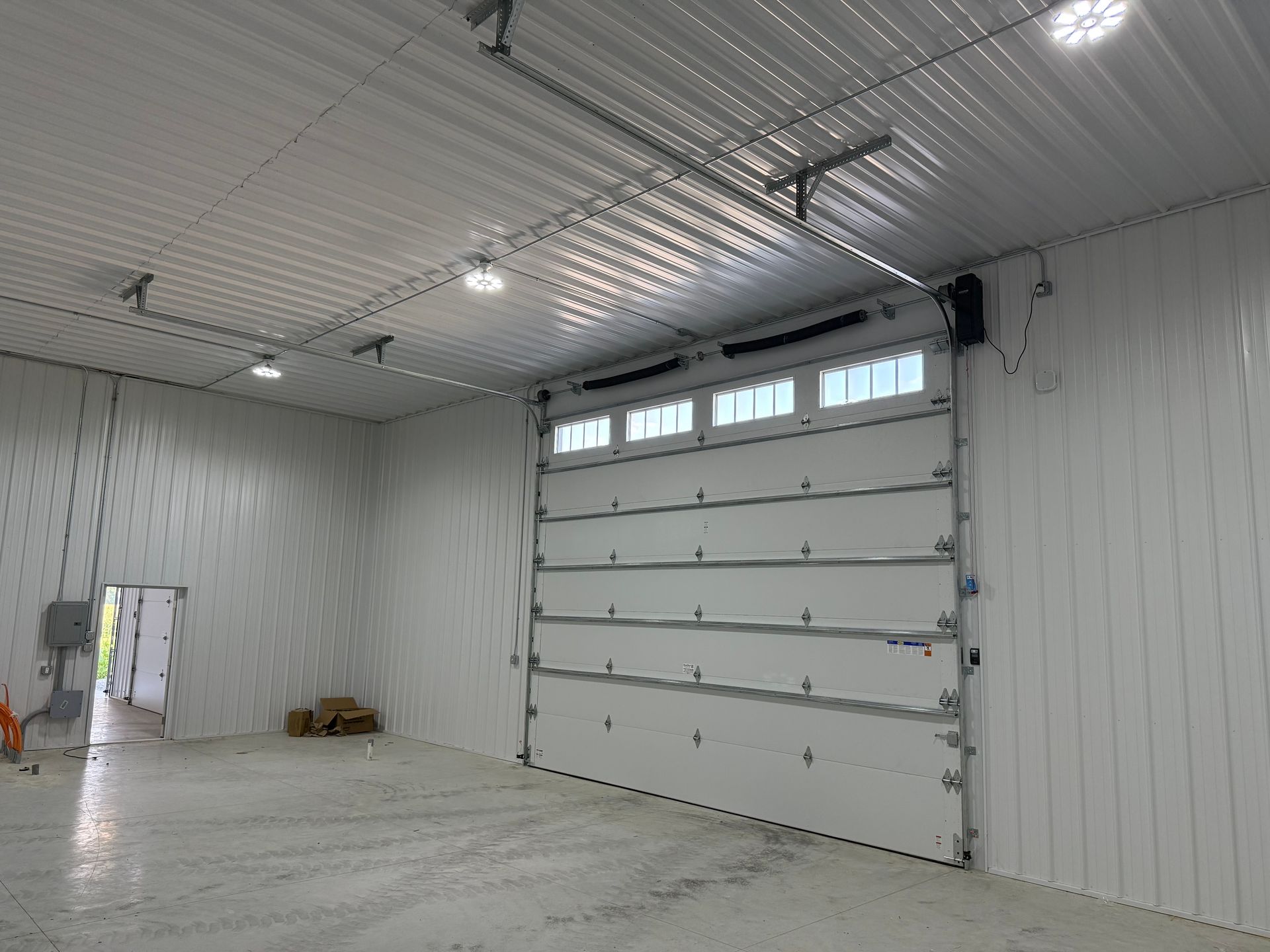 Empty industrial warehouse interior with white walls, concrete floor, overhead lights, and a large garage door