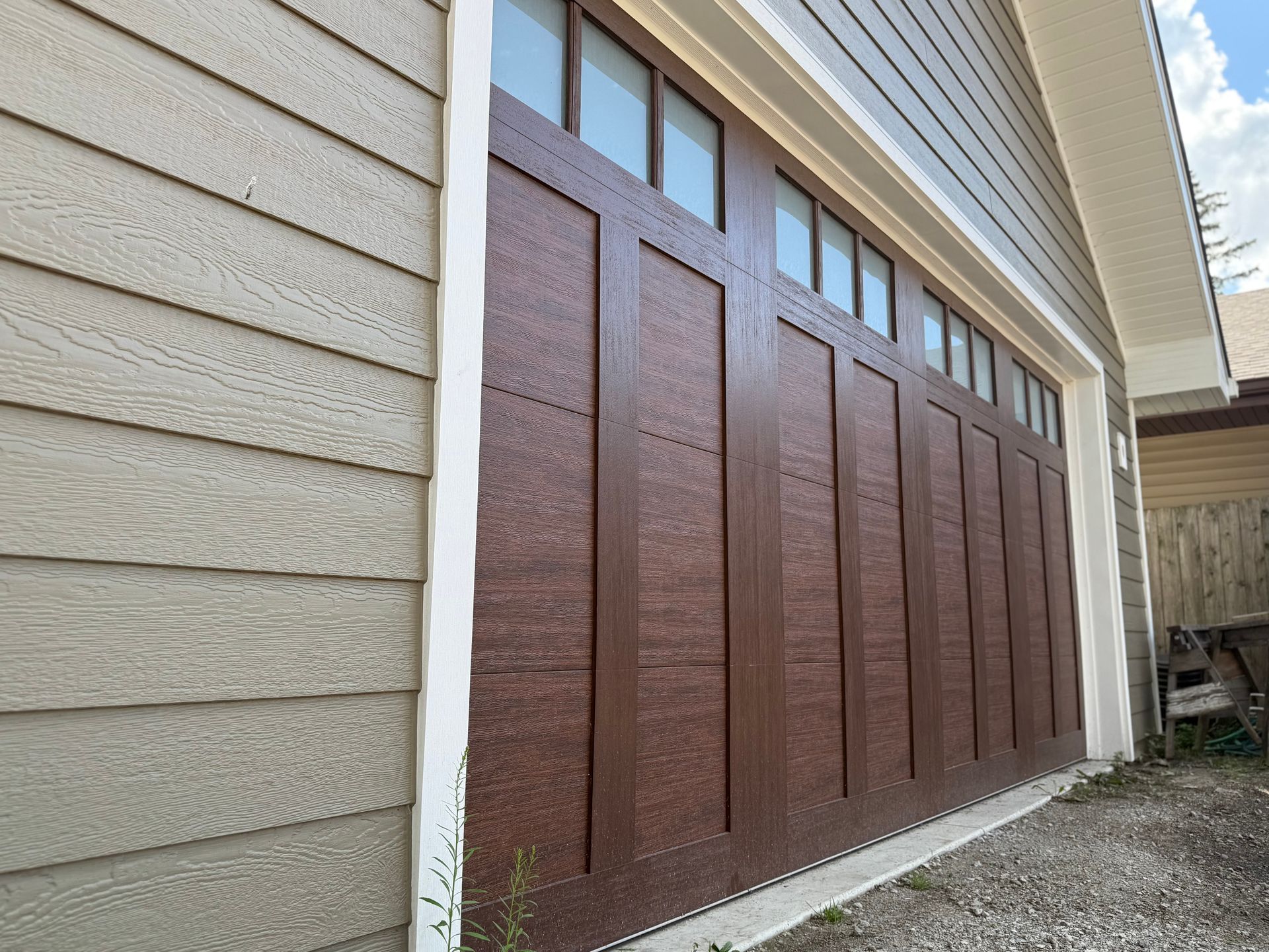 Brown garage doors along the side of a beige house in a narrow outdoor driveway.