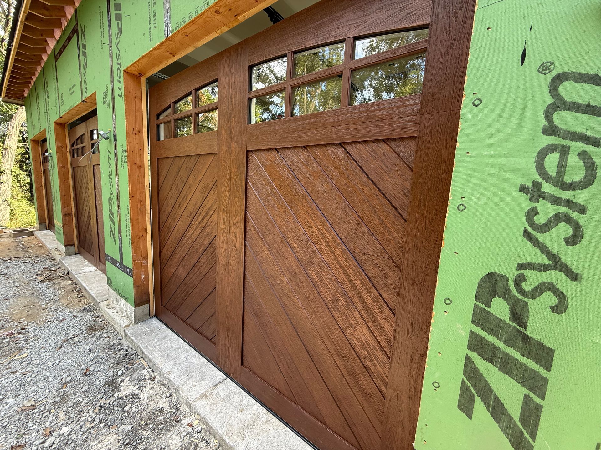 Wooden garage doors in a green stucco building exterior with gravel driveway and trees reflected in windows