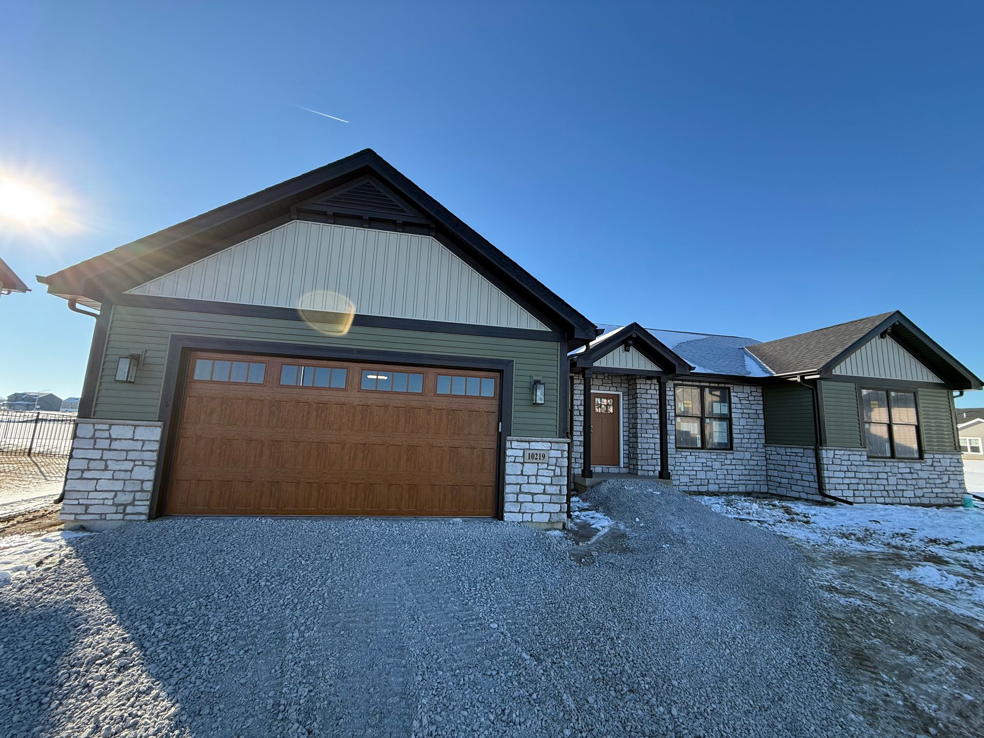 Single-story house with stone and wood exterior, attached garage, and snowy driveway under a bright blue sky