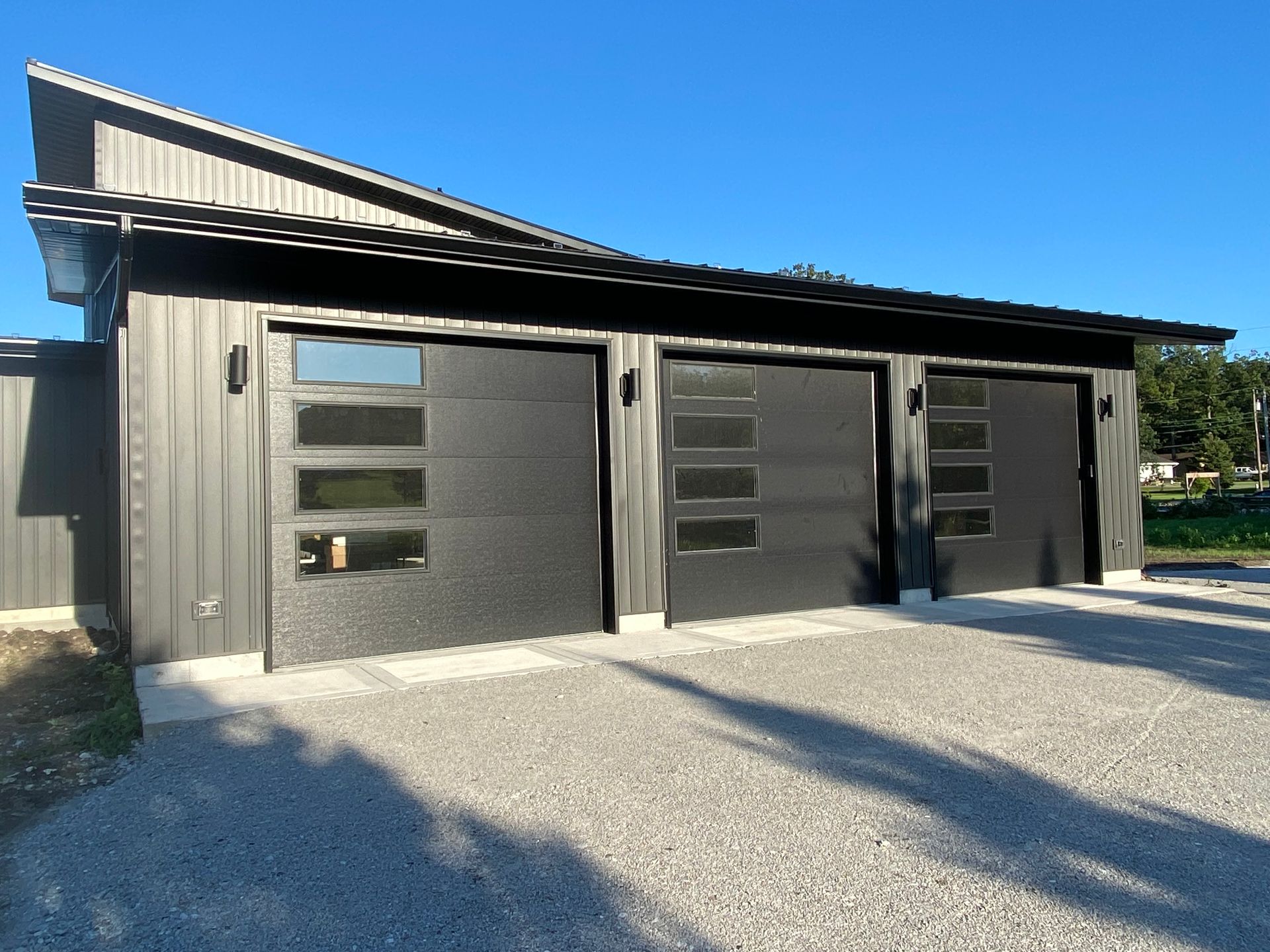 Modern black-and-gray three-car garage with metal siding and gravel driveway in bright sunlight