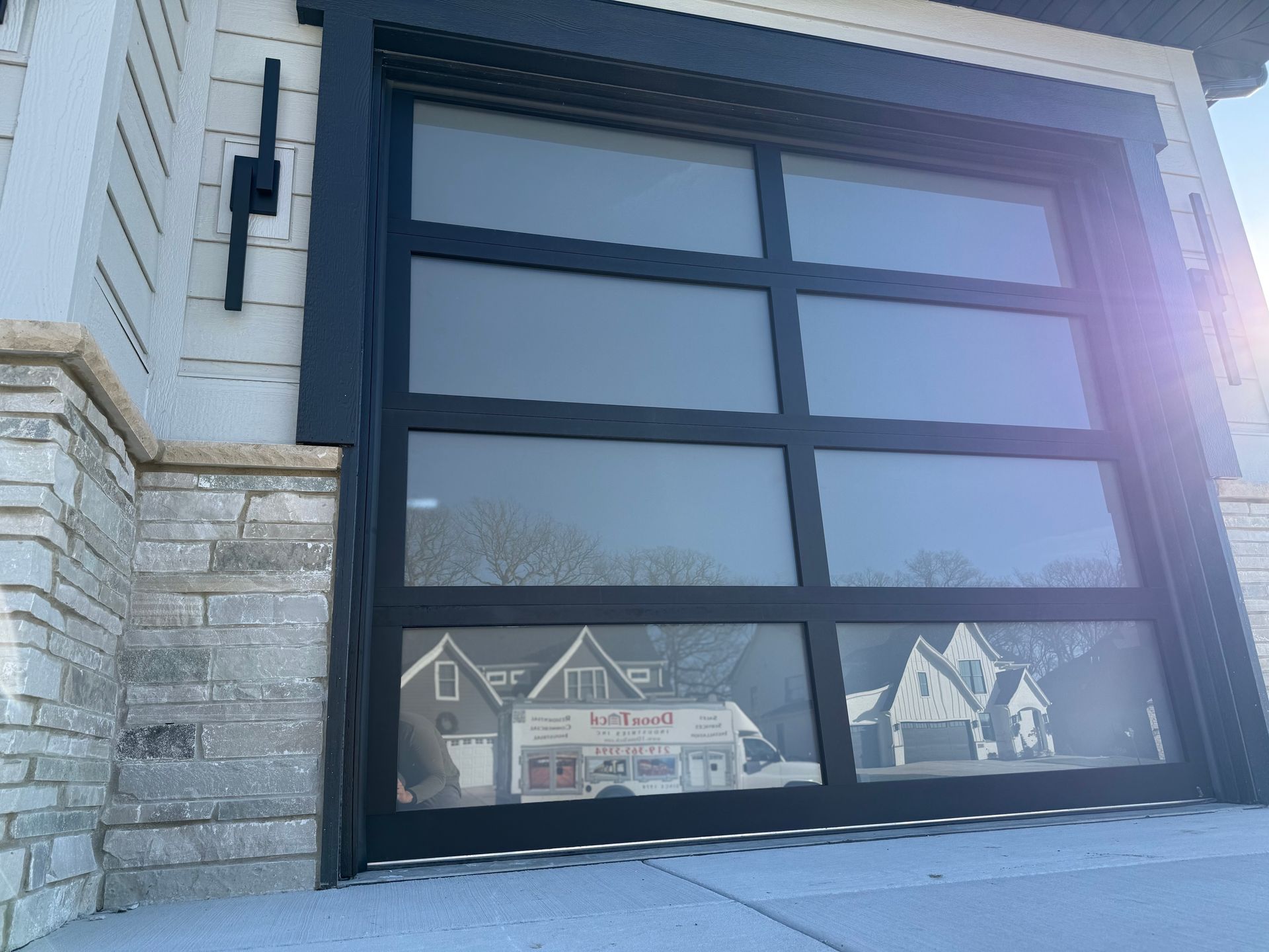 Modern black garage door on a stone-and-siding house, with bright sun glare and reflections of homes.