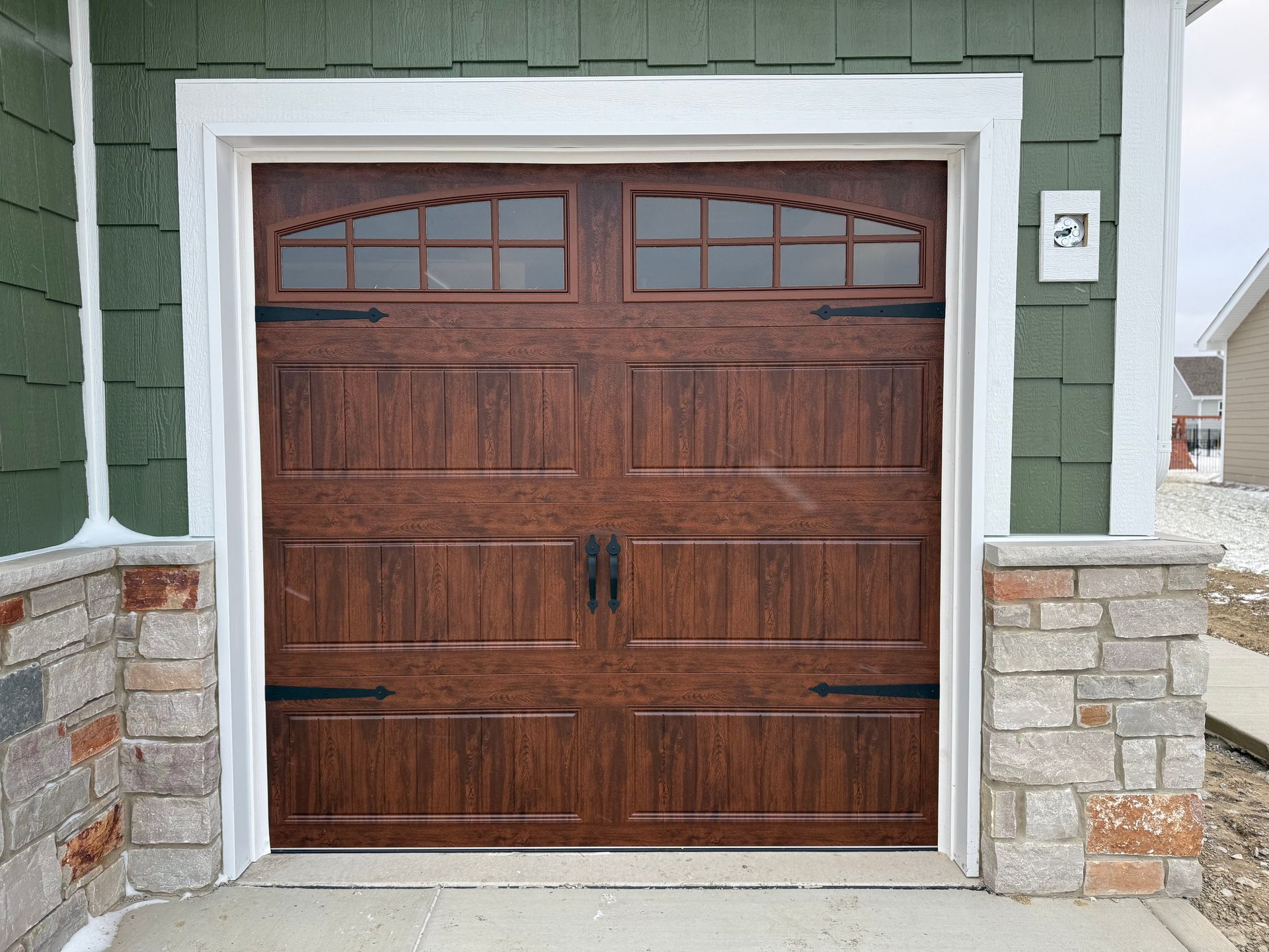 Wooden double garage door with small windows, framed by white trim and stone pillars on a green house.
