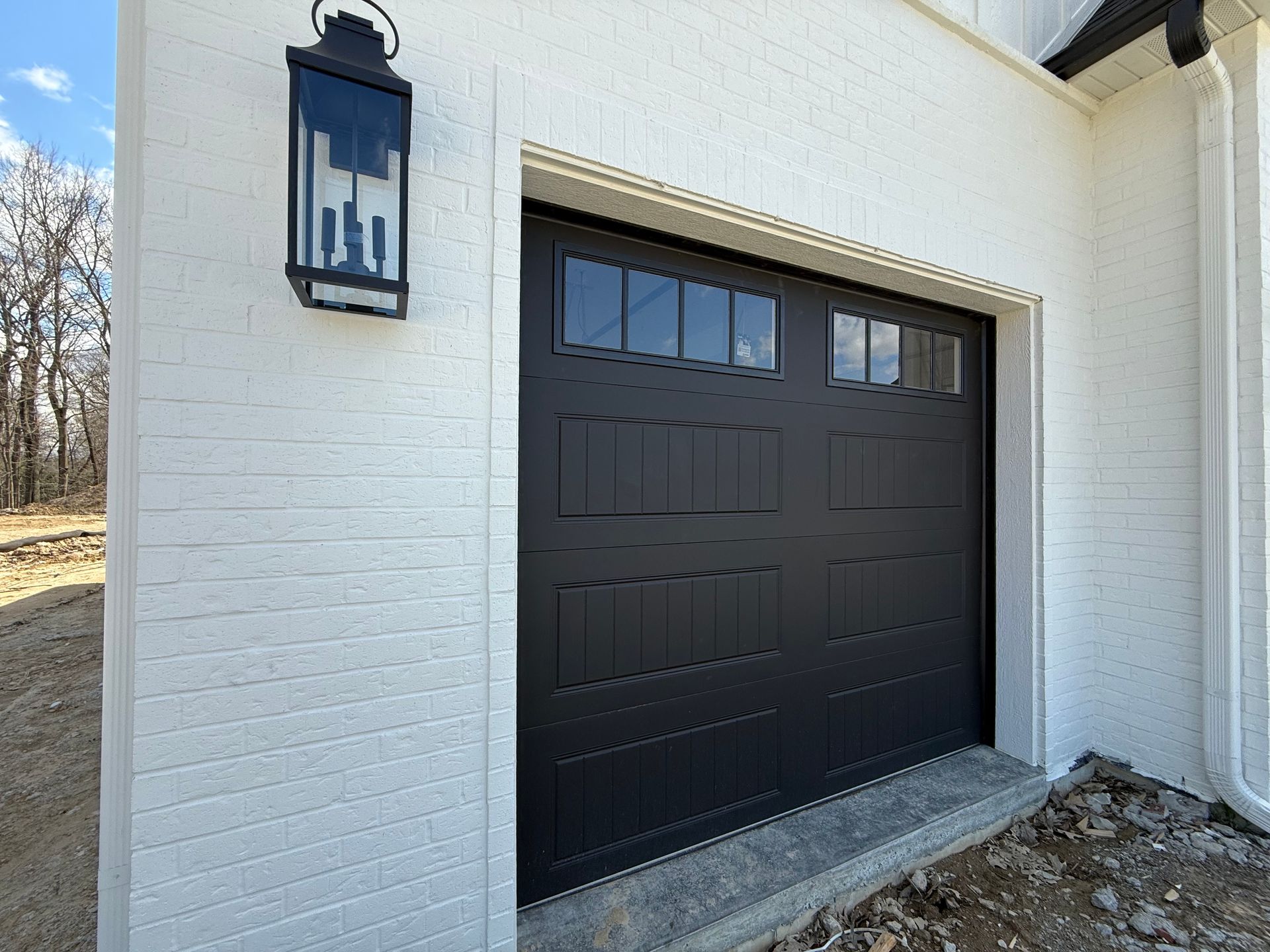 Black garage door on a white brick house exterior with a wall lantern.