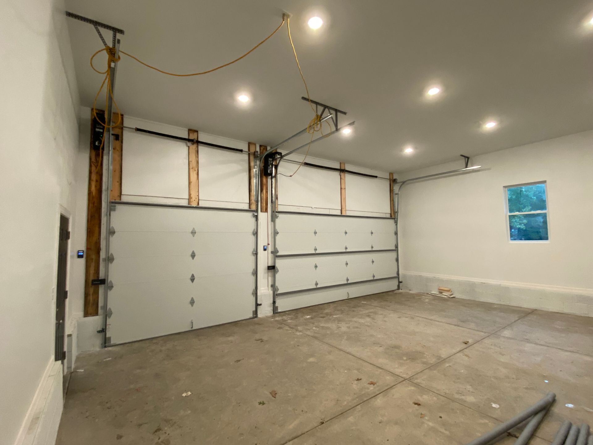 Empty garage interior with white roll-up doors, bare concrete floor, and recessed ceiling lights.