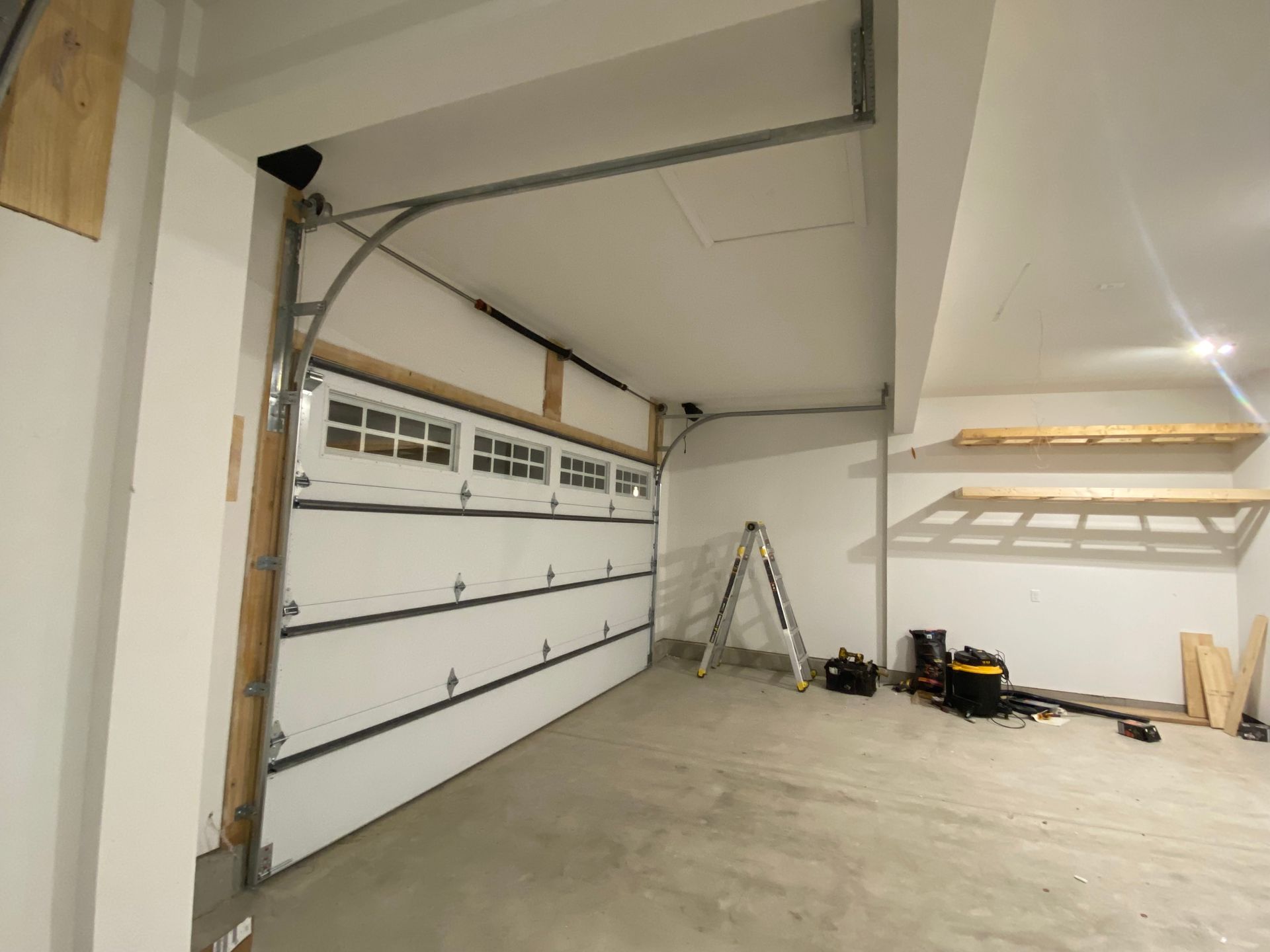 Empty garage interior with white walls, open garage door, ladder, and tools on the floor