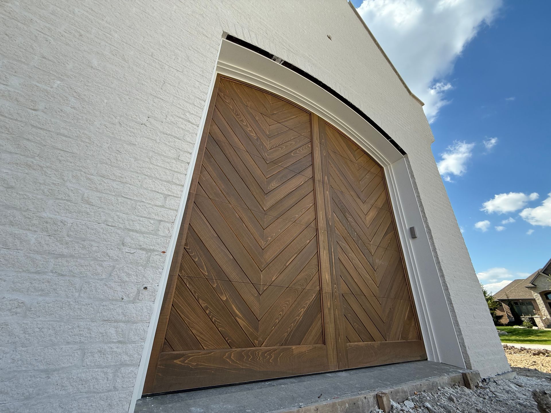 Wooden double doors with herringbone panels set in a curved stucco wall under a blue sky.