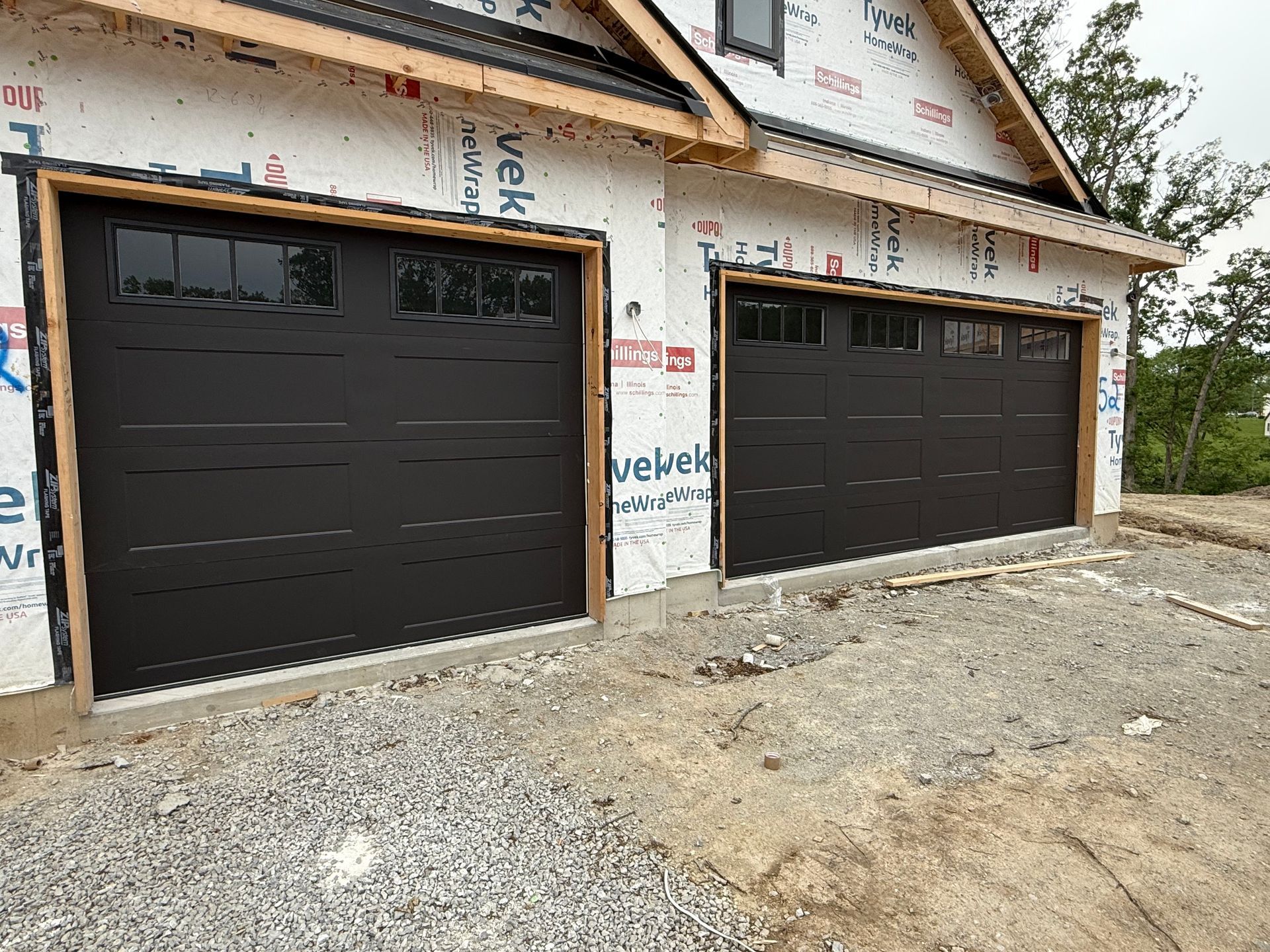 Two black garage doors on a house under construction, with unfinished siding and gravel driveway.