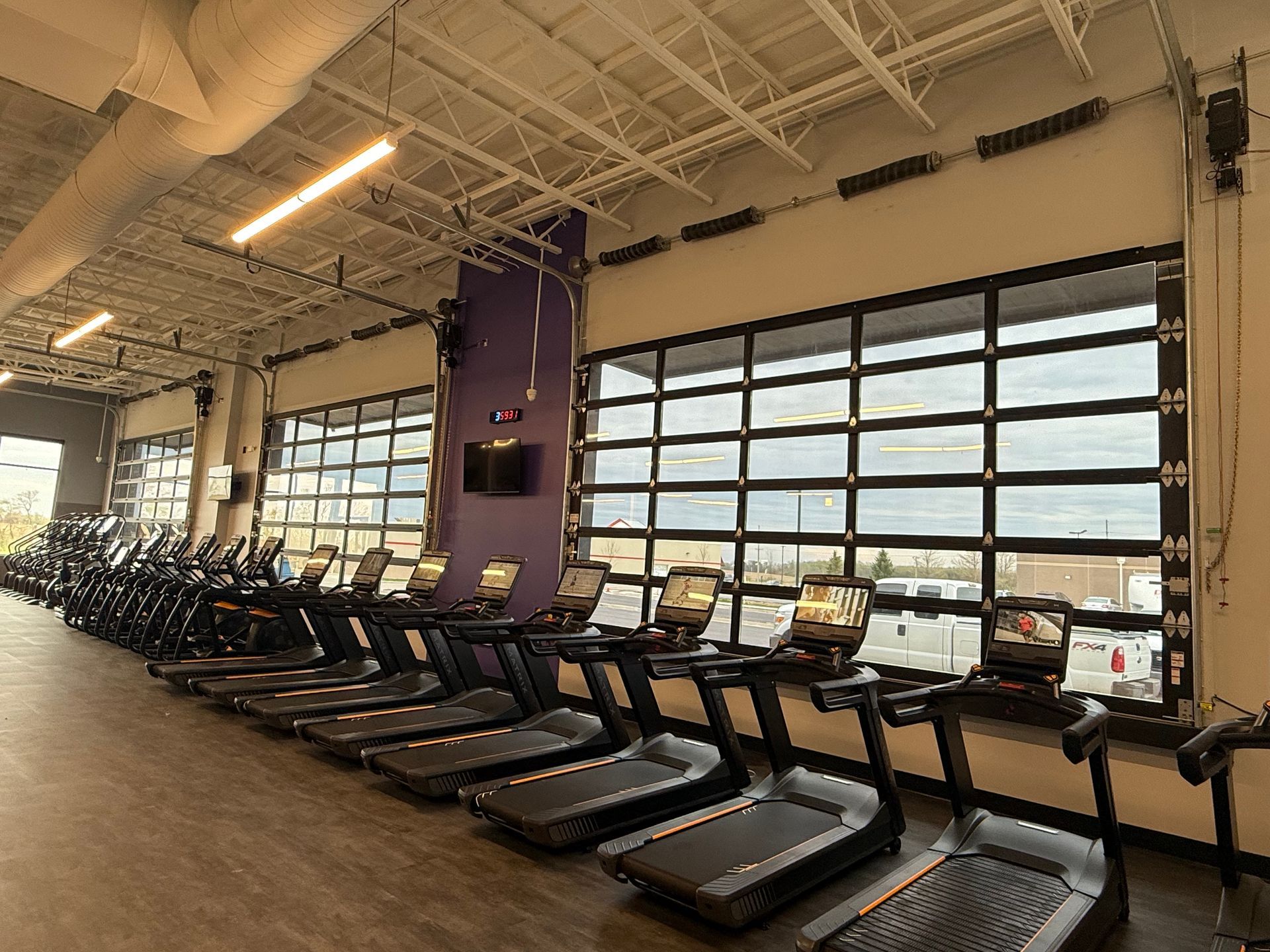 Rows of treadmills in a bright gym with large windows and a purple pillar