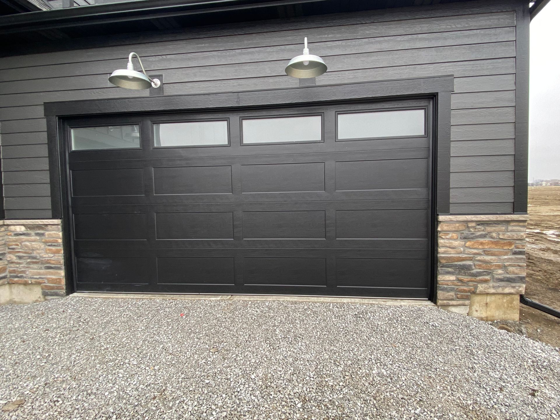 Modern black garage door on a gray stone house with two wall lights and gravel driveway