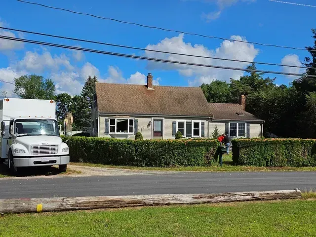 White moving truck parked in front of a tan house with a green hedge. Person trimming the hedge. Bright blue sky.