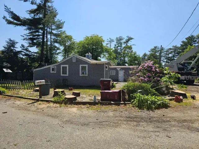 Small gray house with cut tree stumps in front, pink flowers, and a crane visible on a sunny day.
