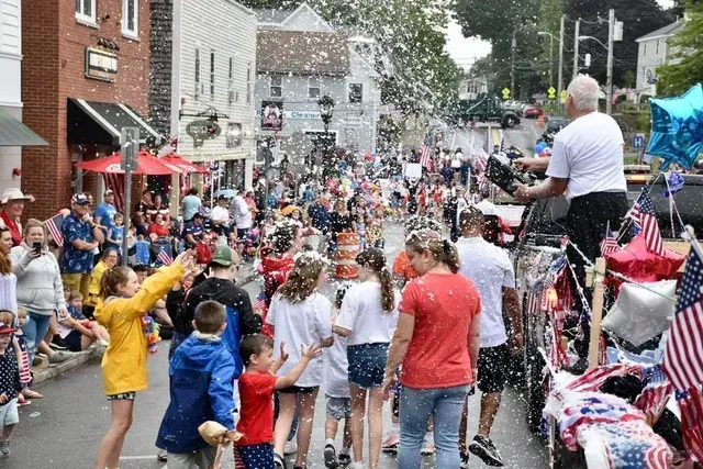 People watch a parade with confetti in a town. Flags and buildings are visible.