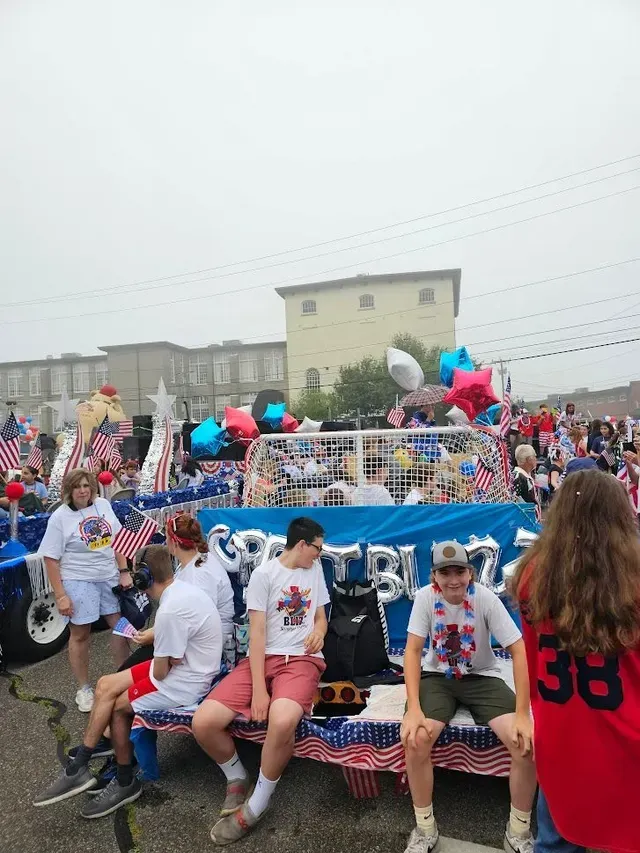 Float decorated with stars, balloons, and flags; people sit/stand on it in a parade.