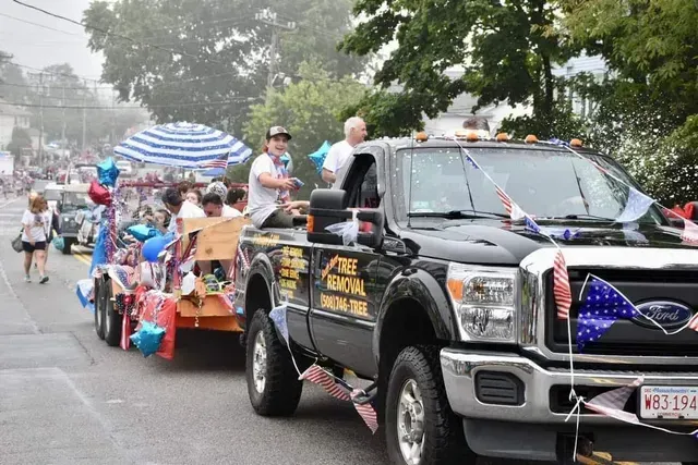 Black truck pulling a trailer in a parade, decorated with flags and balloons. People are on the truck and trailer.