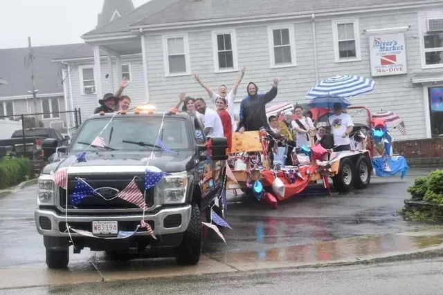 A truck pulling a decorated trailer in a parade; people waving under umbrellas, in front of a building.