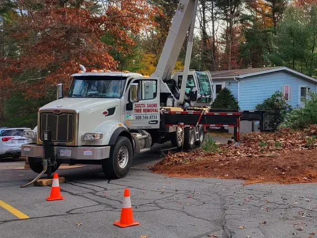 A white crane truck parked on a paved lot with two orange traffic cones in the foreground and a blue house in the back.