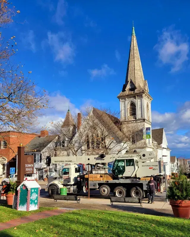 Crane next to a historic church with a tall steeple on a sunny day. A decorated box and potted plants are in the foreground.