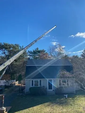 A crane extending above a house with a dark roof against a blue sky, trees on both sides.