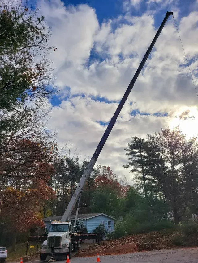 A crane extends upwards, trees and a house in the background under a cloudy sky.