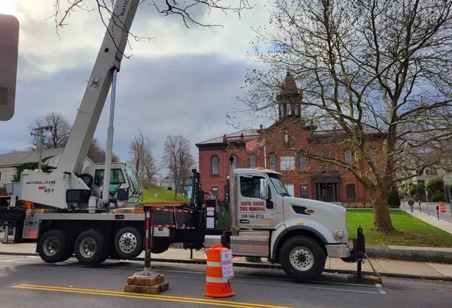 A crane truck parked on a street near a brick building. The sky is cloudy. An orange traffic cone is present.