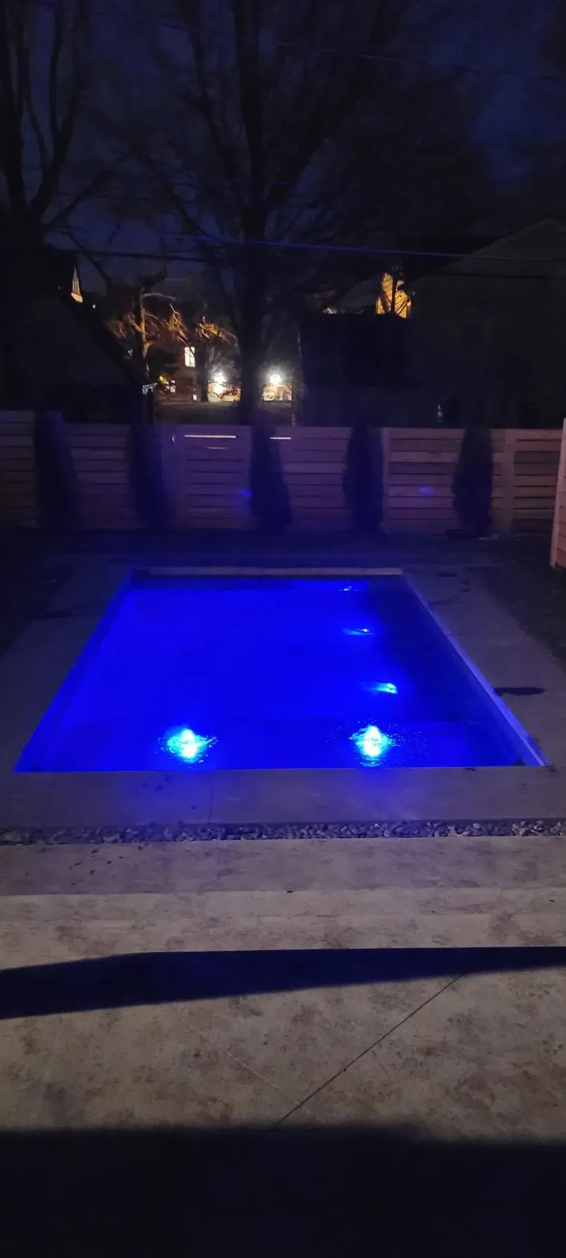 A rectangular pool illuminated with blue lights at night, surrounded by concrete and a wooden fence.